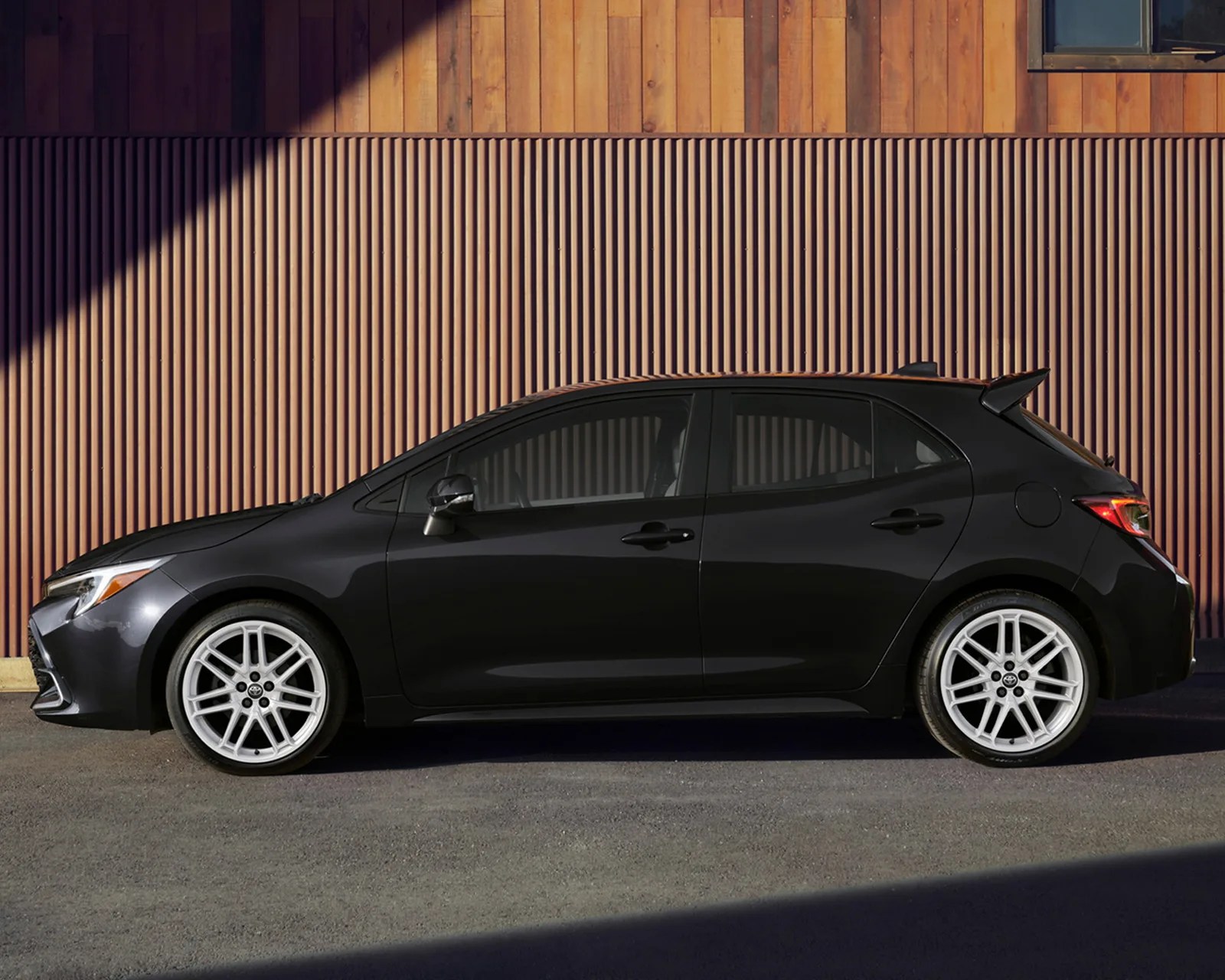Black hatchback car with white alloy wheels parked in front of a wooden and corrugated metal wall.