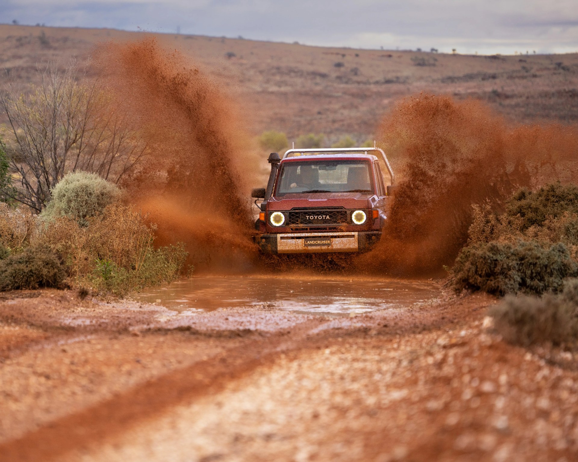 Red Toyota Land Cruiser splashing muddy water on a dirt road in a dry, shrub-filled landscape.