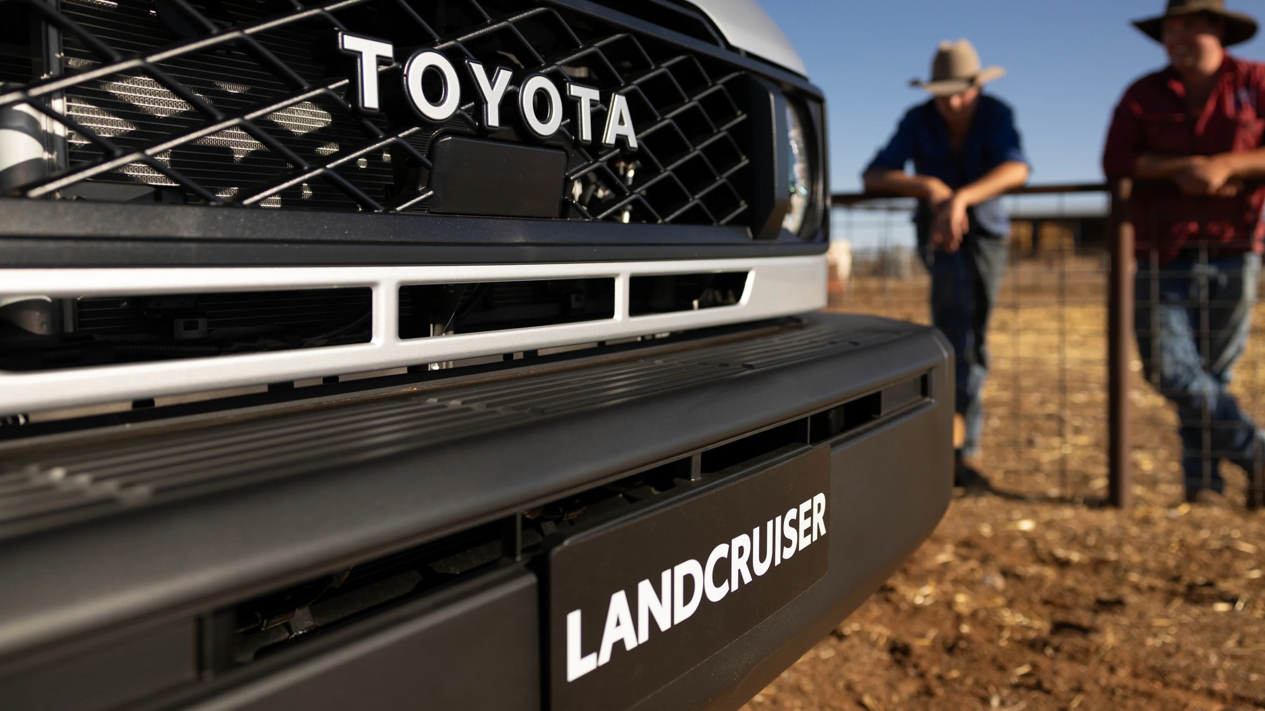 Close-up of a silver Toyota Land Cruiser front grille and bumper with two people in cowboy hats leaning on a fence in the background.