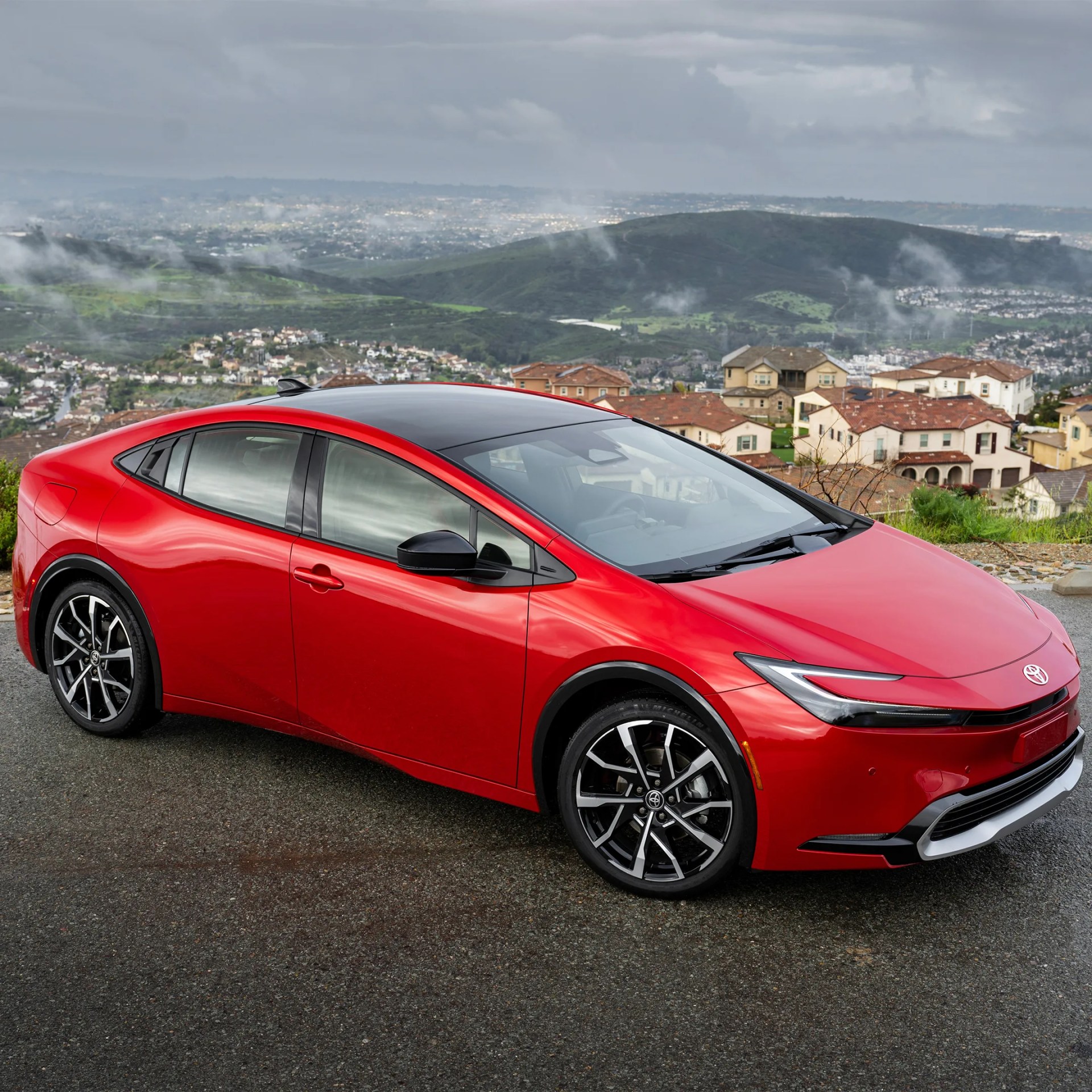 Red Toyota sedan with black roof and alloy wheels parked on a wet road with houses and hills in the background.