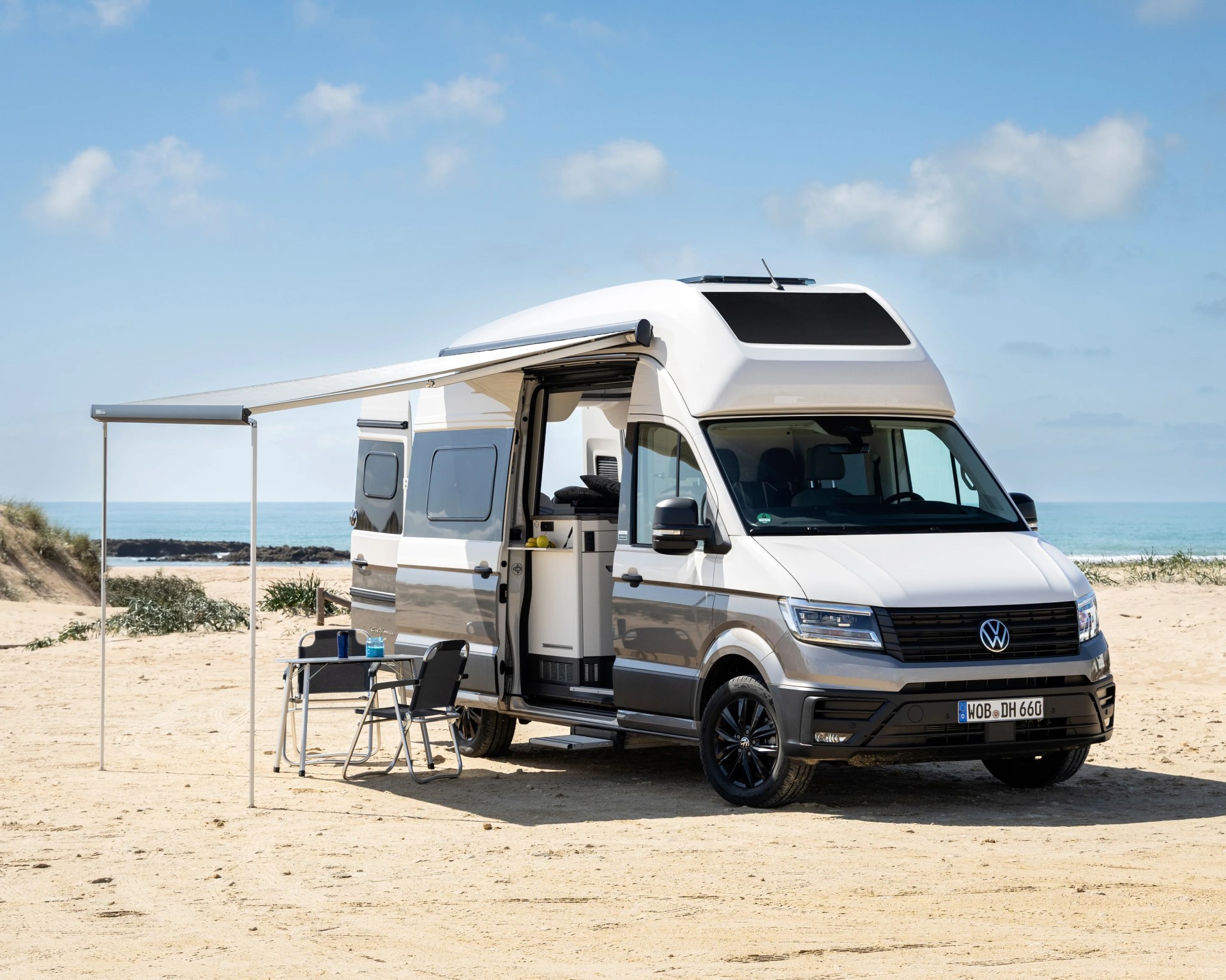 White and gray Volkswagen camper van with extended awning and outdoor table and chairs on a sandy beach.