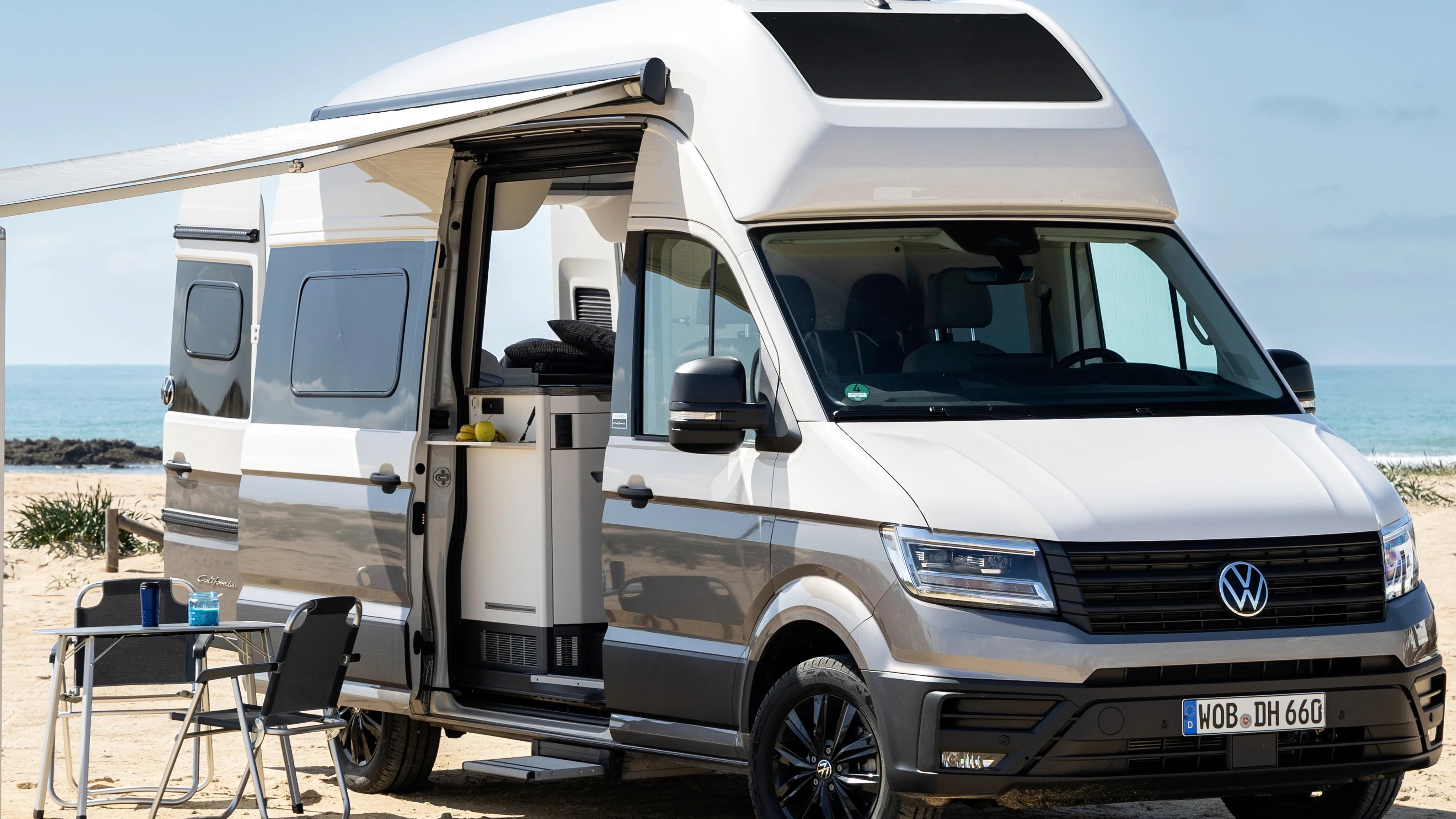 White and gray Volkswagen camper van with open side door and awning on a sandy beach.