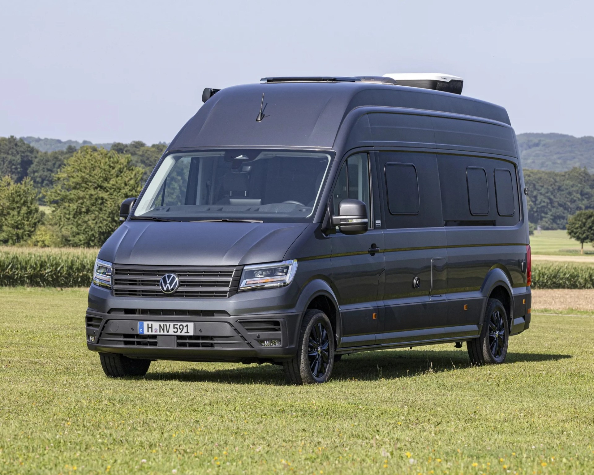 Dark gray Volkswagen camper van with black wheels parked on grass with trees in the background.