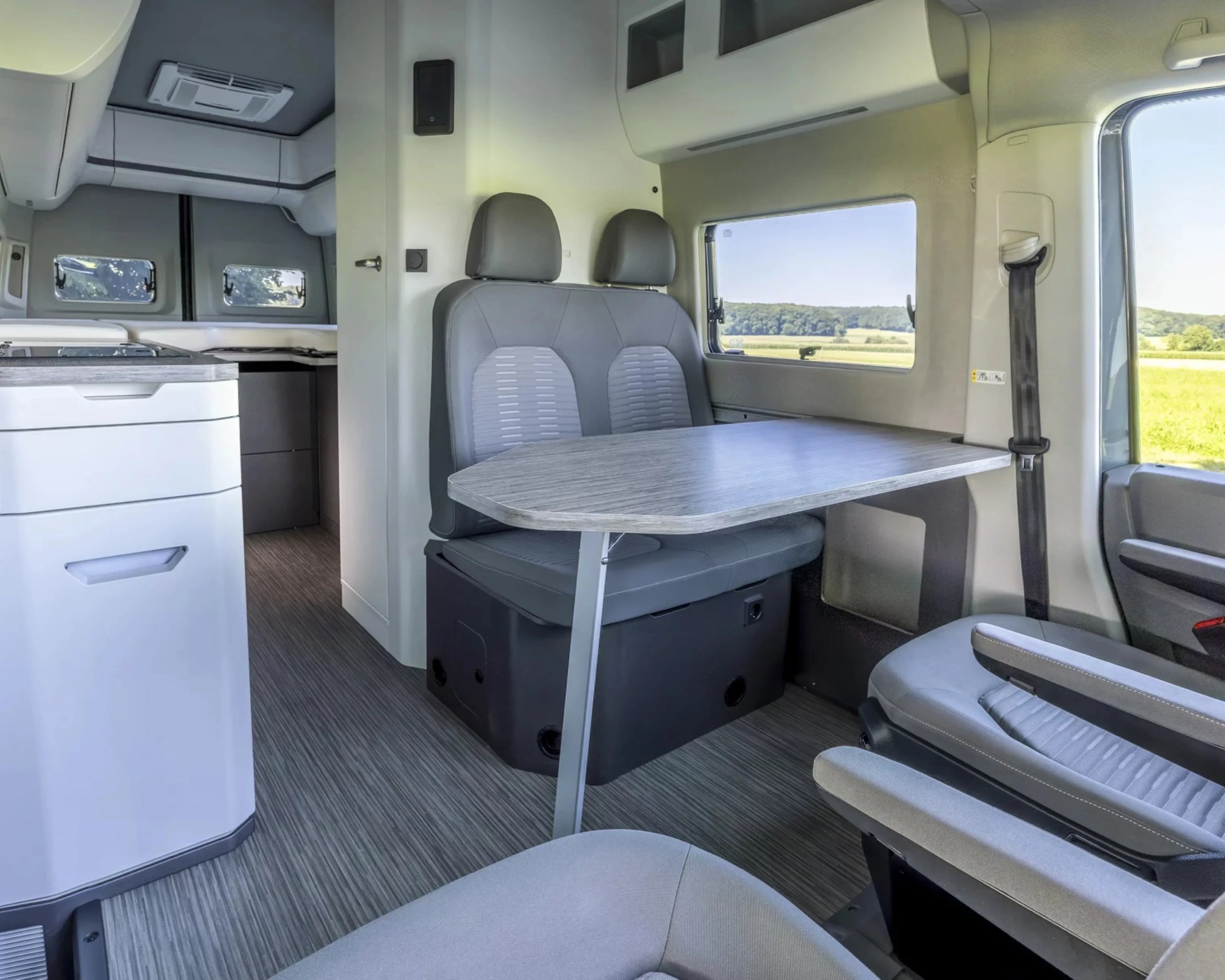 Interior of a camper van with gray cushioned bench seating, a foldable wooden table, and a kitchenette in the background.
