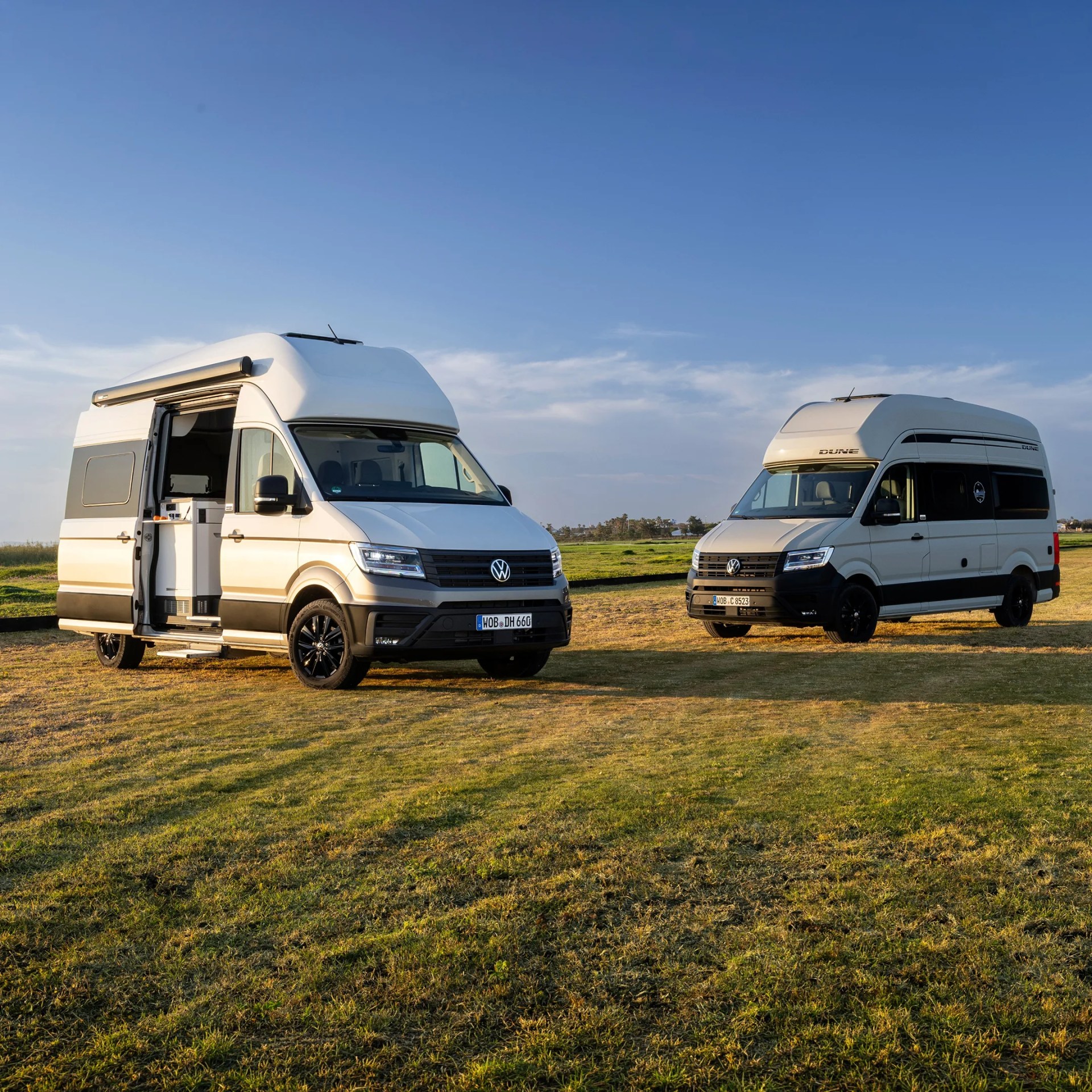 Two white Volkswagen camper vans parked on grass under a clear blue sky, one with an open side door showing the interior.