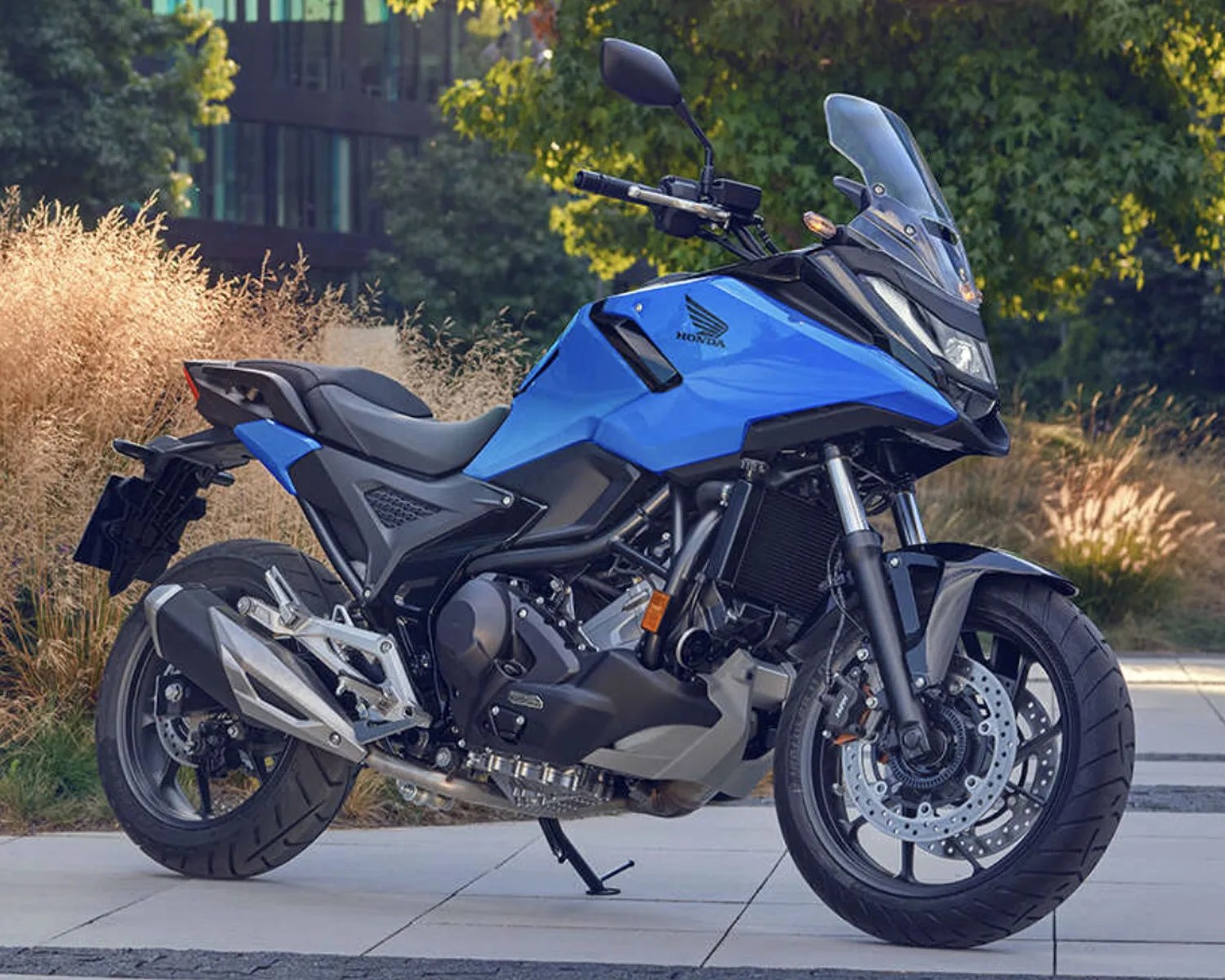 Blue and black Honda adventure motorcycle parked on a sidewalk with trees and grass in the background.