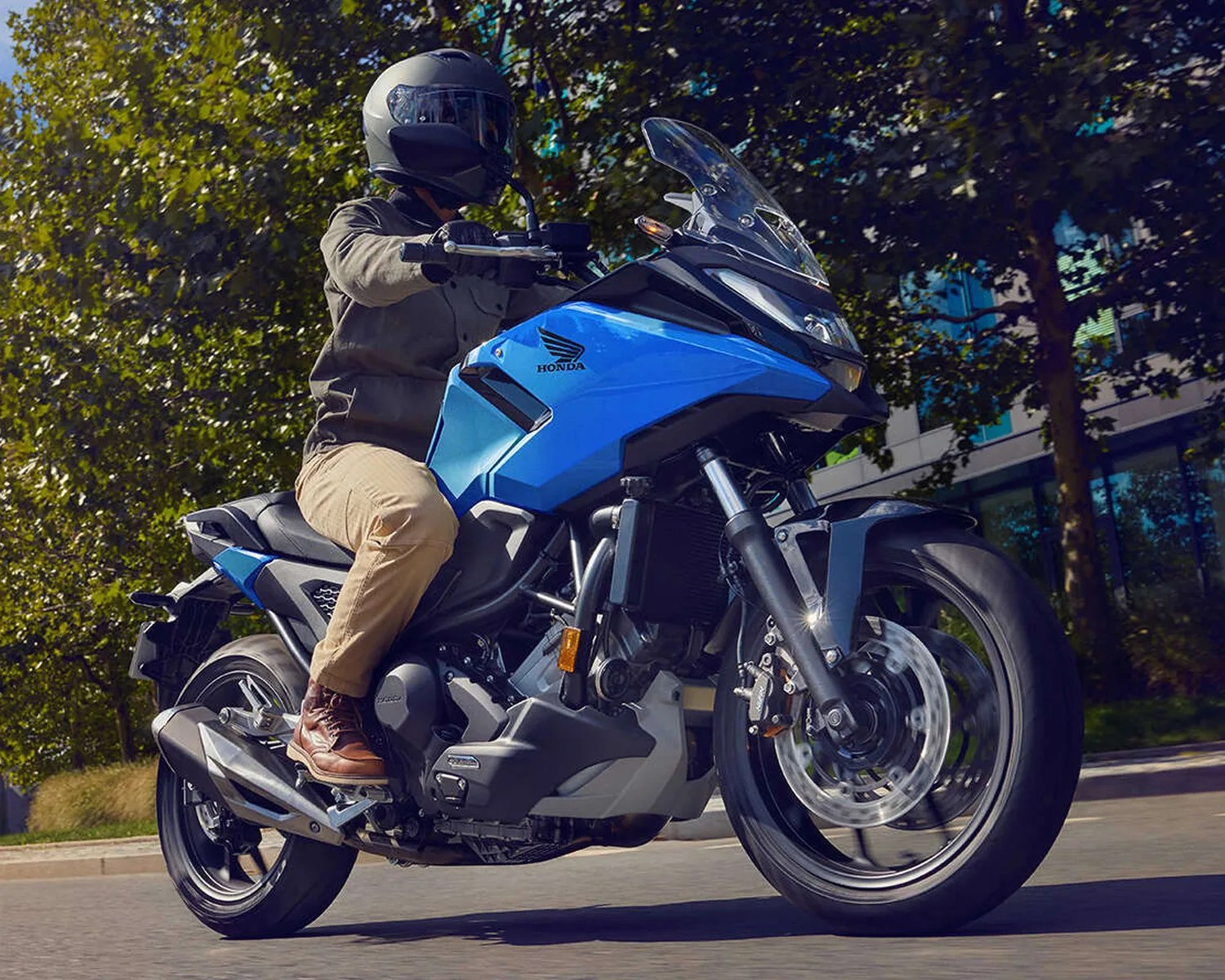 Person wearing a black helmet riding a blue Honda motorcycle on a street with trees and buildings in the background.