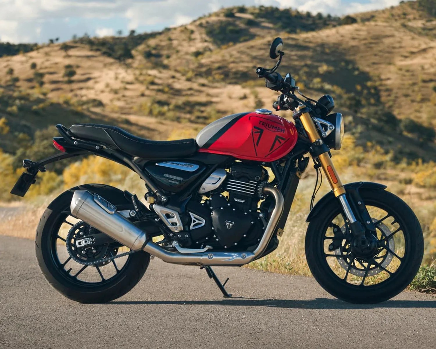 Red and silver Triumph motorcycle with black seat and gold front forks parked on a road with hills in the background.