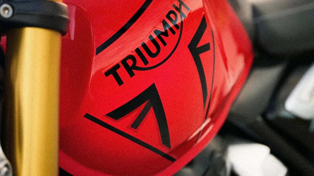 Close-up of a red motorcycle fuel tank with black "TRIUMPH" logo and gold front fork.