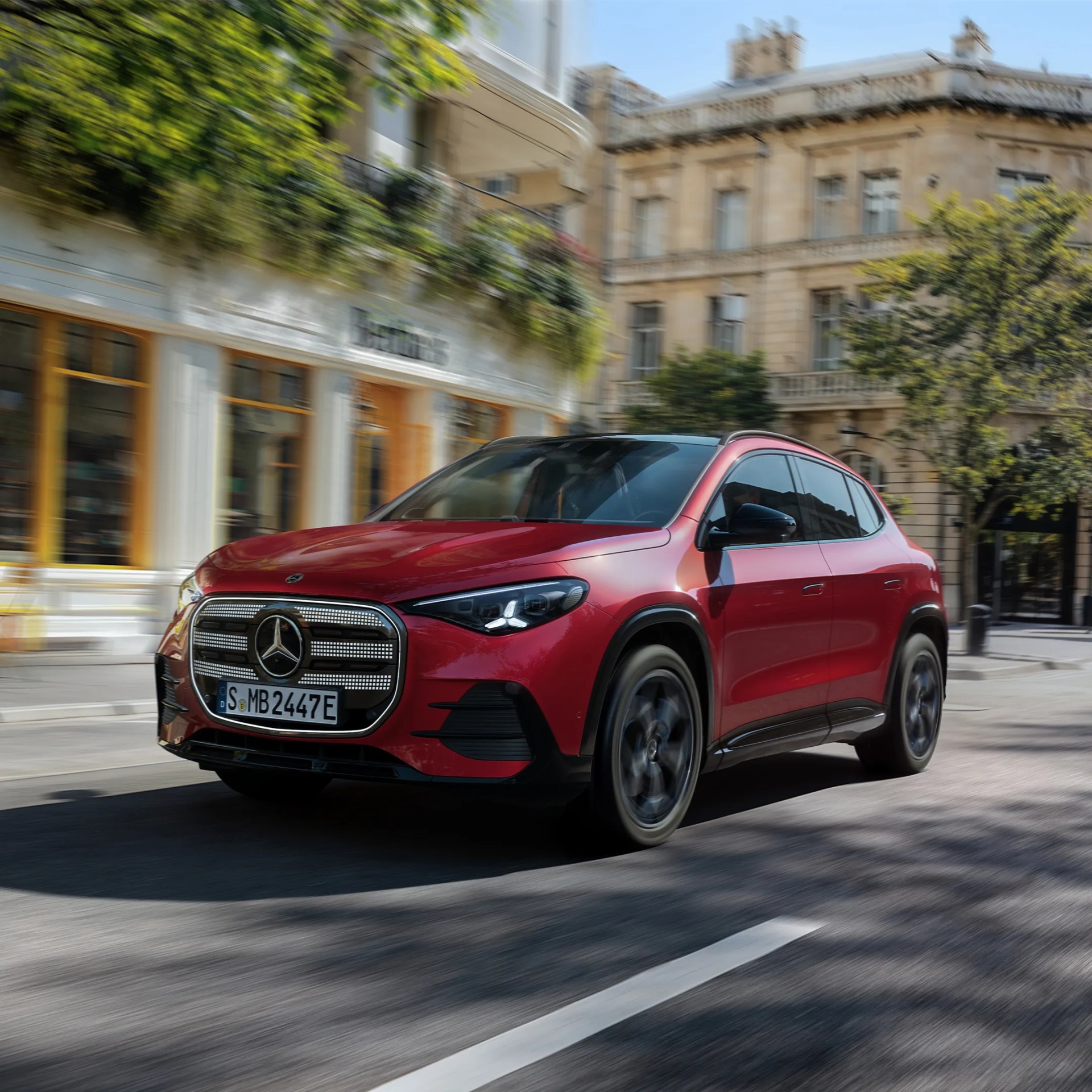 Red Mercedes-Benz SUV driving on a city street with historic buildings and greenery in the background.