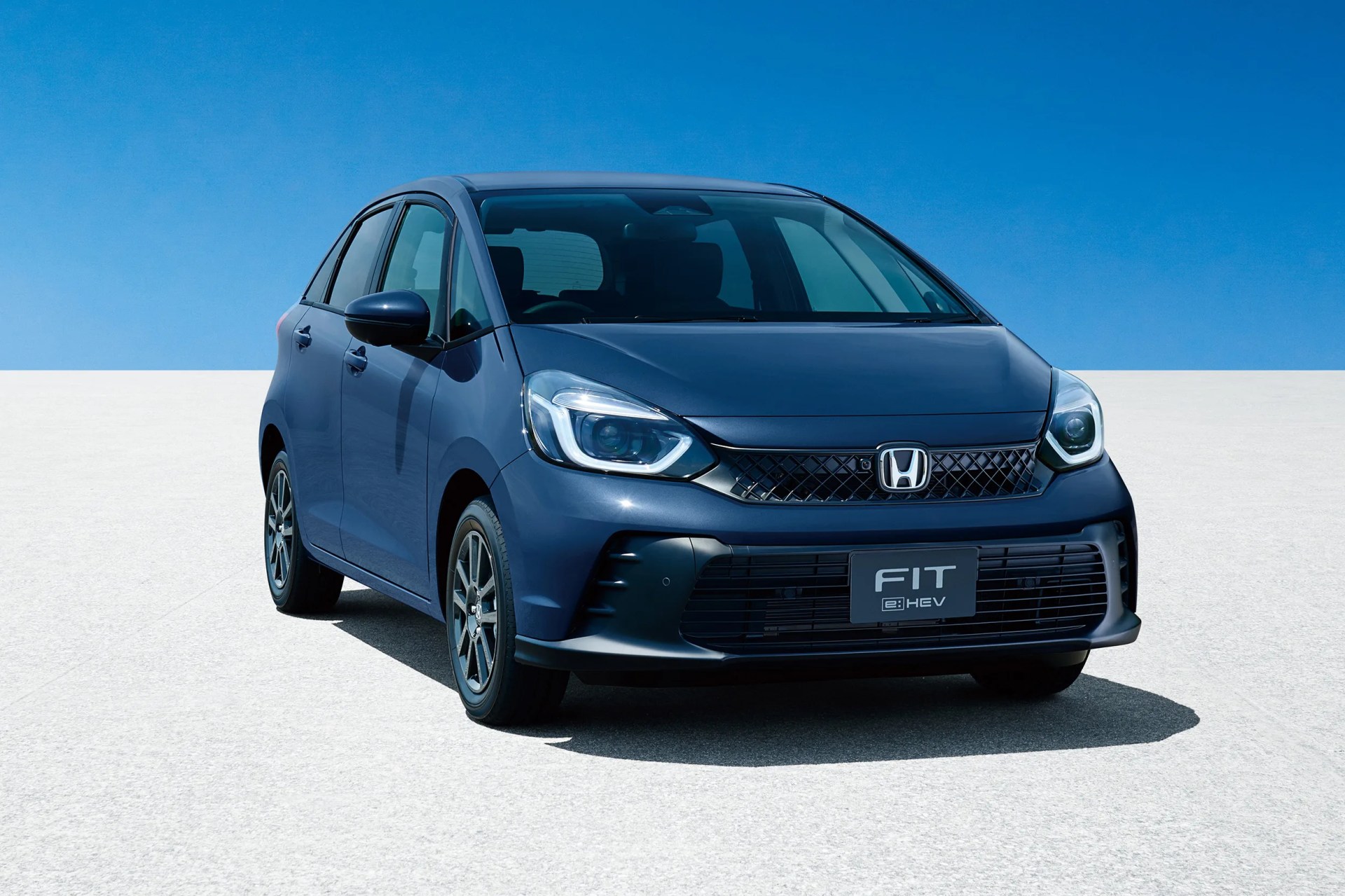 Blue Honda Fit e:HEV compact car front view on a light concrete surface under clear blue sky.