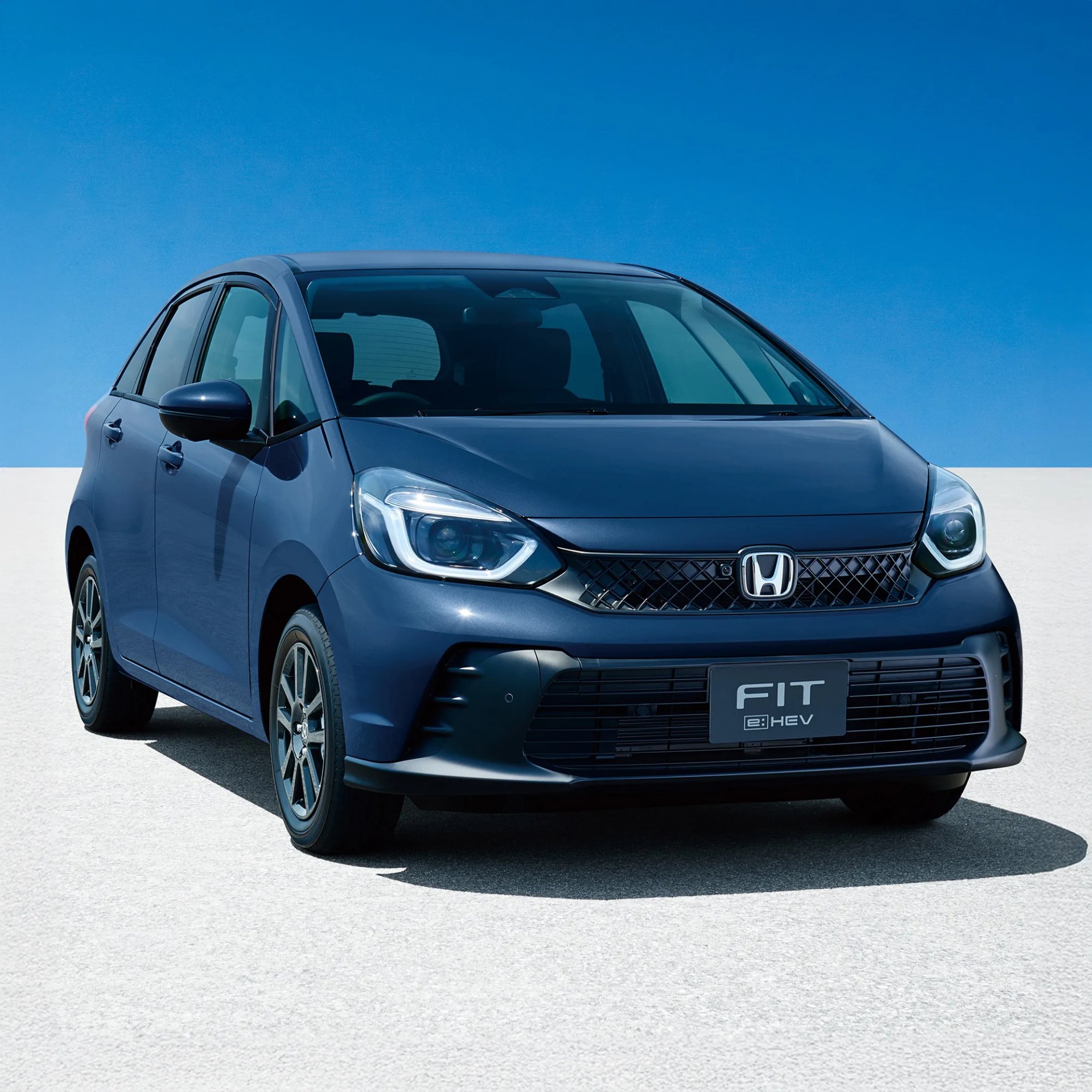 Blue Honda Fit e:HEV compact car front view on a light gray surface under a clear blue sky.