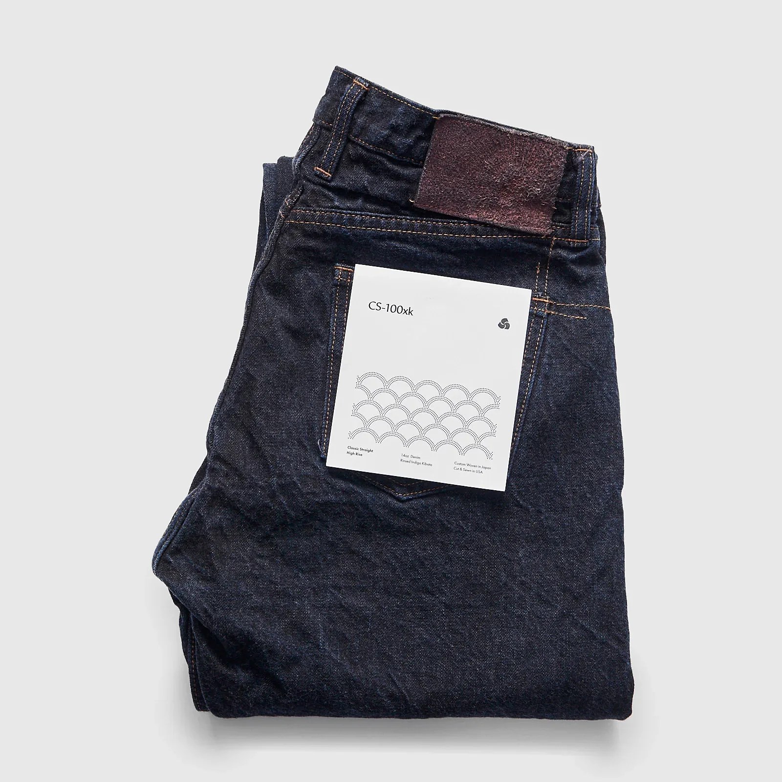 Folded dark indigo denim jeans with a white label on the back pocket reading "CS-100xk".