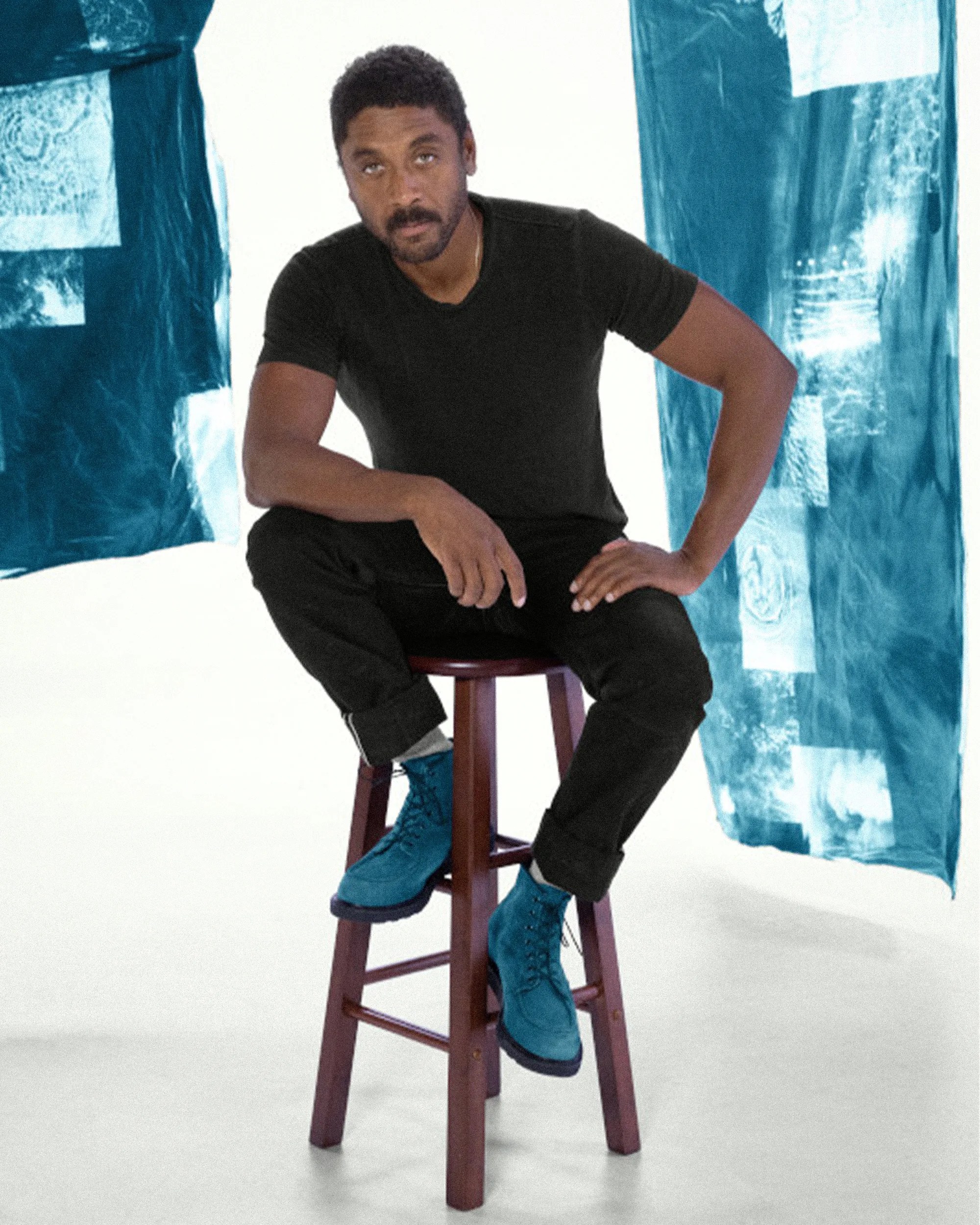 Man wearing black t-shirt and pants sitting on a wooden stool with blue lace-up boots against a white and blue fabric backdrop.