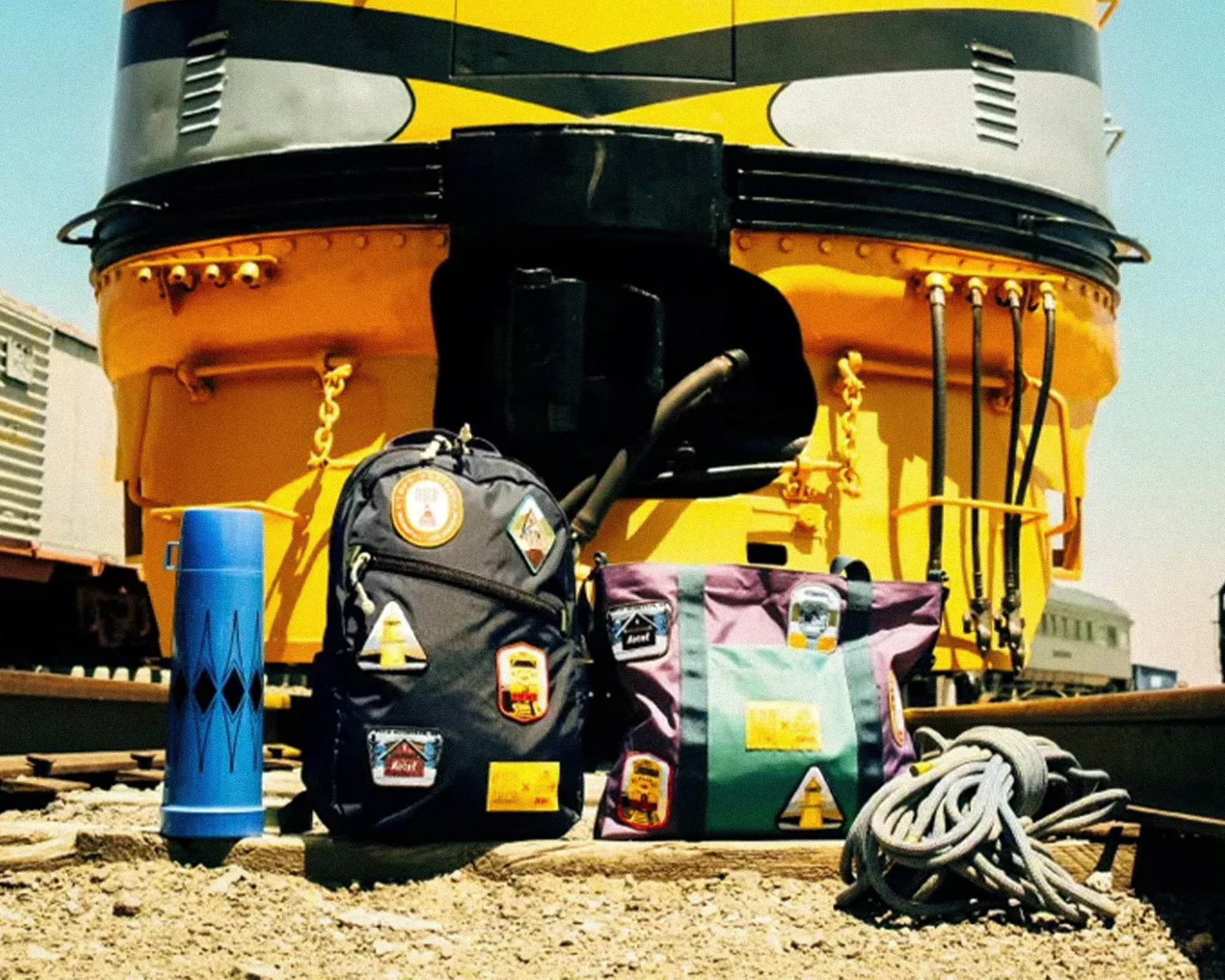 Two backpacks with patches, a rolled rope, and a blue cylindrical object in front of a yellow and black train engine.