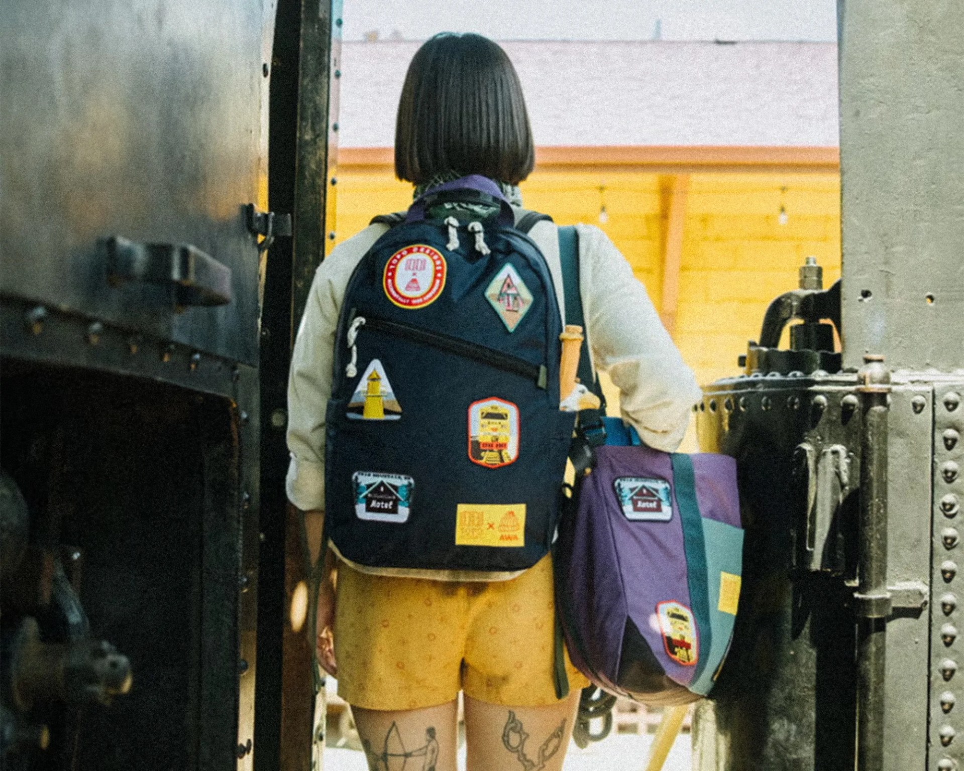 Person with short dark hair wearing a black backpack with patches and carrying a purple and teal bag, standing between metal structures.