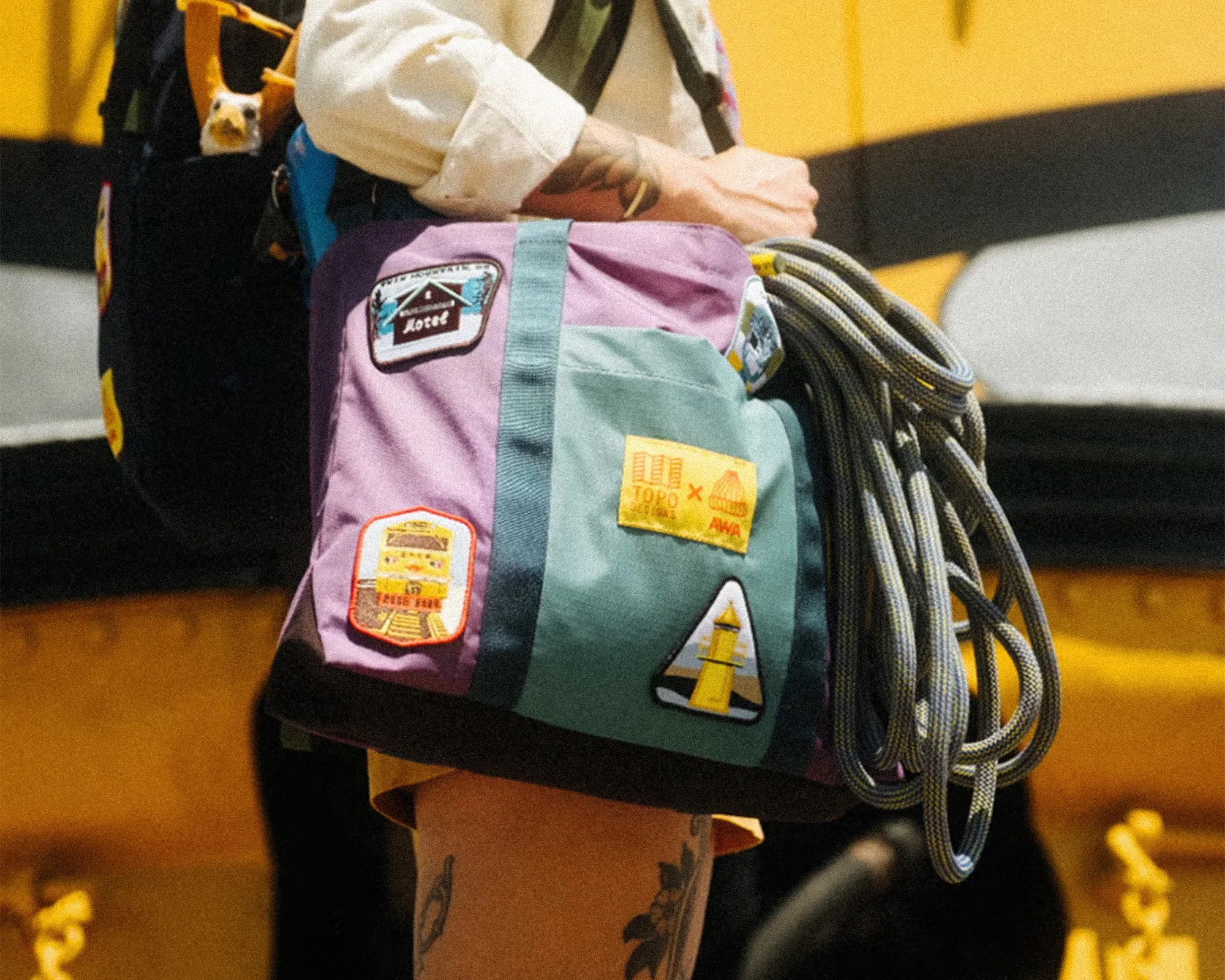 Purple and green climbing chalk bag with patches and a coiled climbing rope attached, worn by a person.