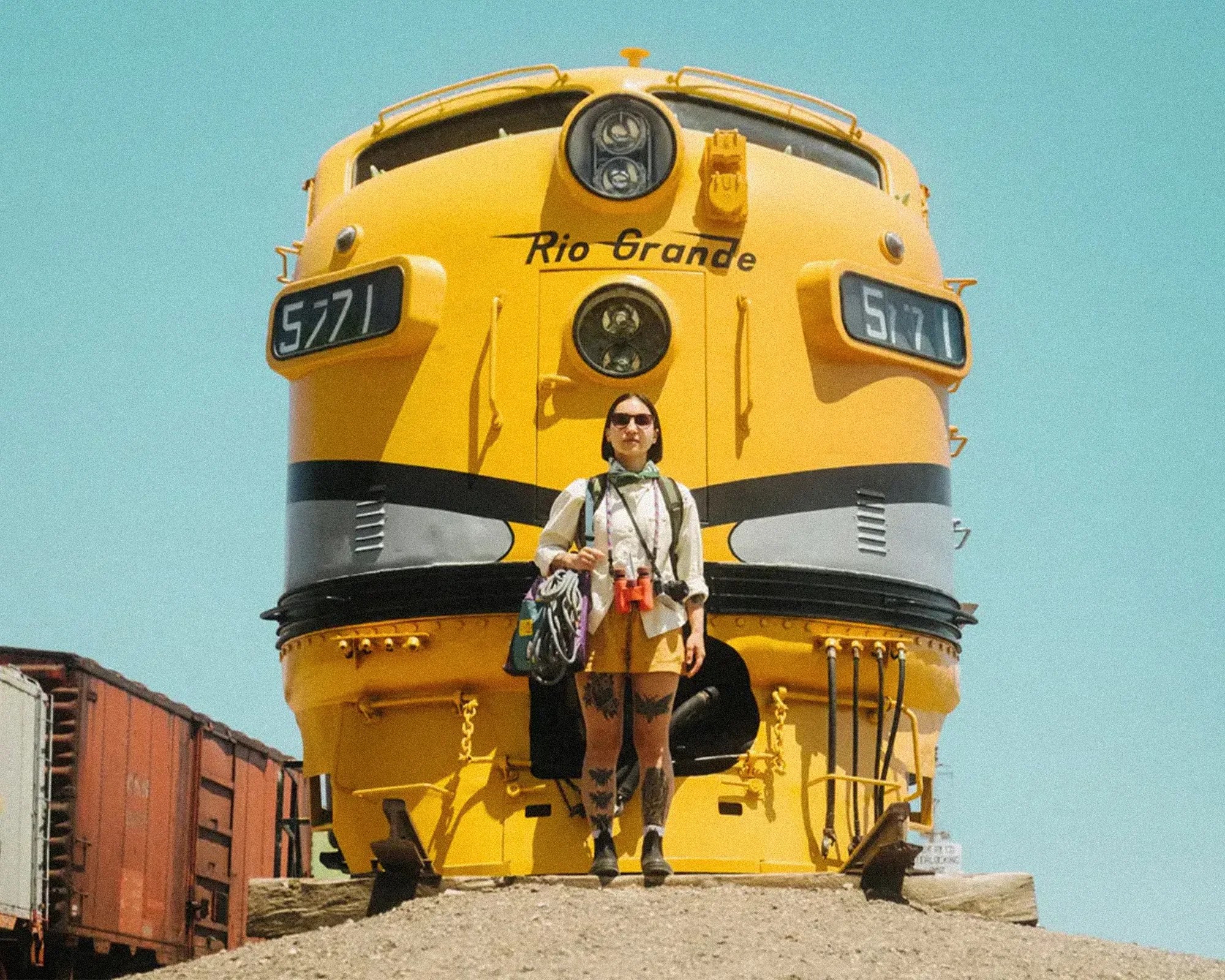 Person with sunglasses and binoculars standing in front of a yellow Rio Grande train engine numbered 5771.