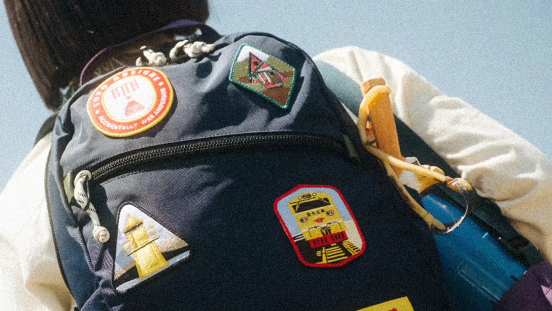 Navy blue backpack with colorful patches and a yellow carabiner attached, worn by a person in a white shirt.