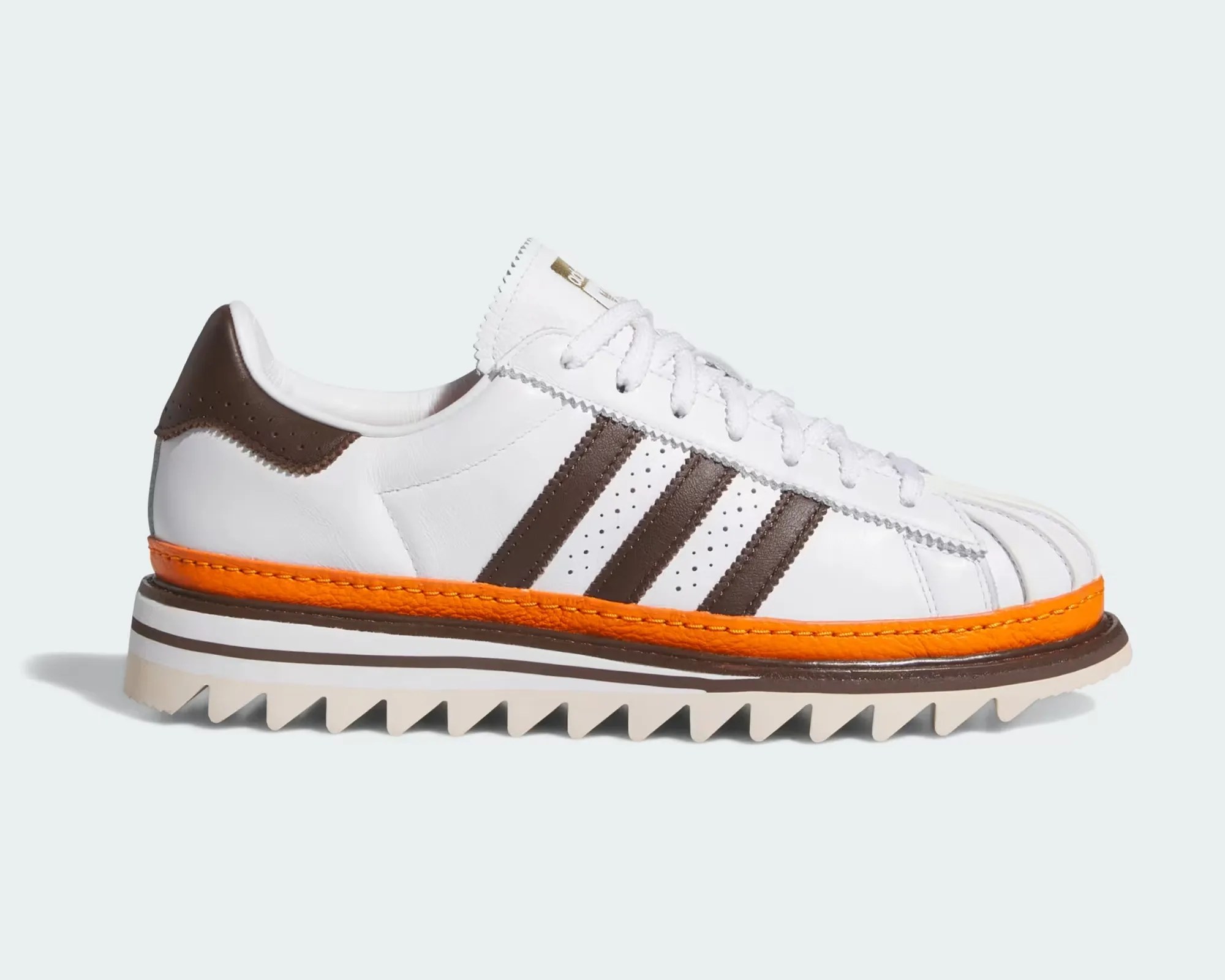 White leather sneaker with brown stripes, brown heel tab, orange midsole, and jagged beige outsole.