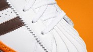 Close-up of a white sneaker with brown leather stripes and white laces on an orange background.