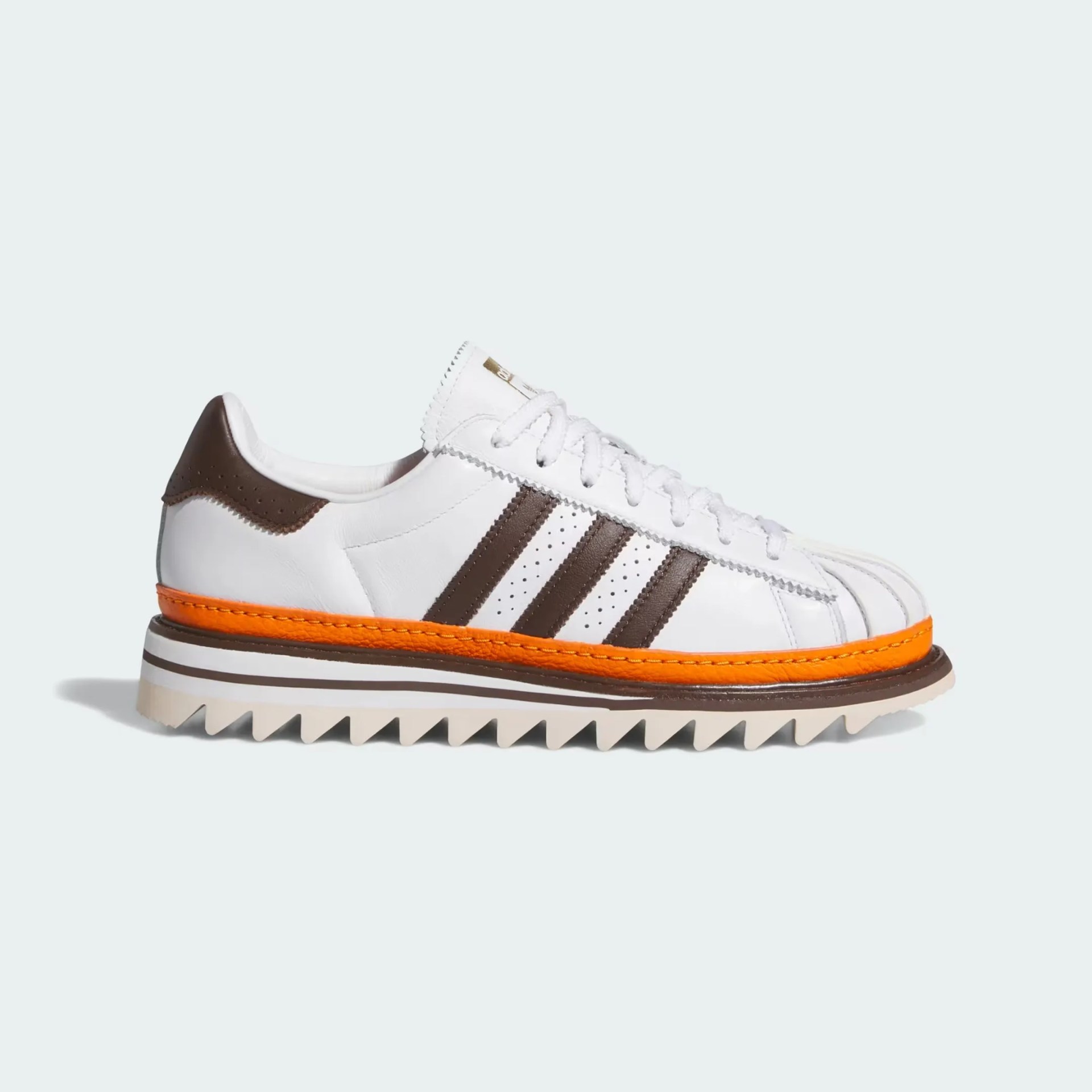 White leather sneaker with brown stripes, brown heel tab, and orange midsole on a jagged white outsole.