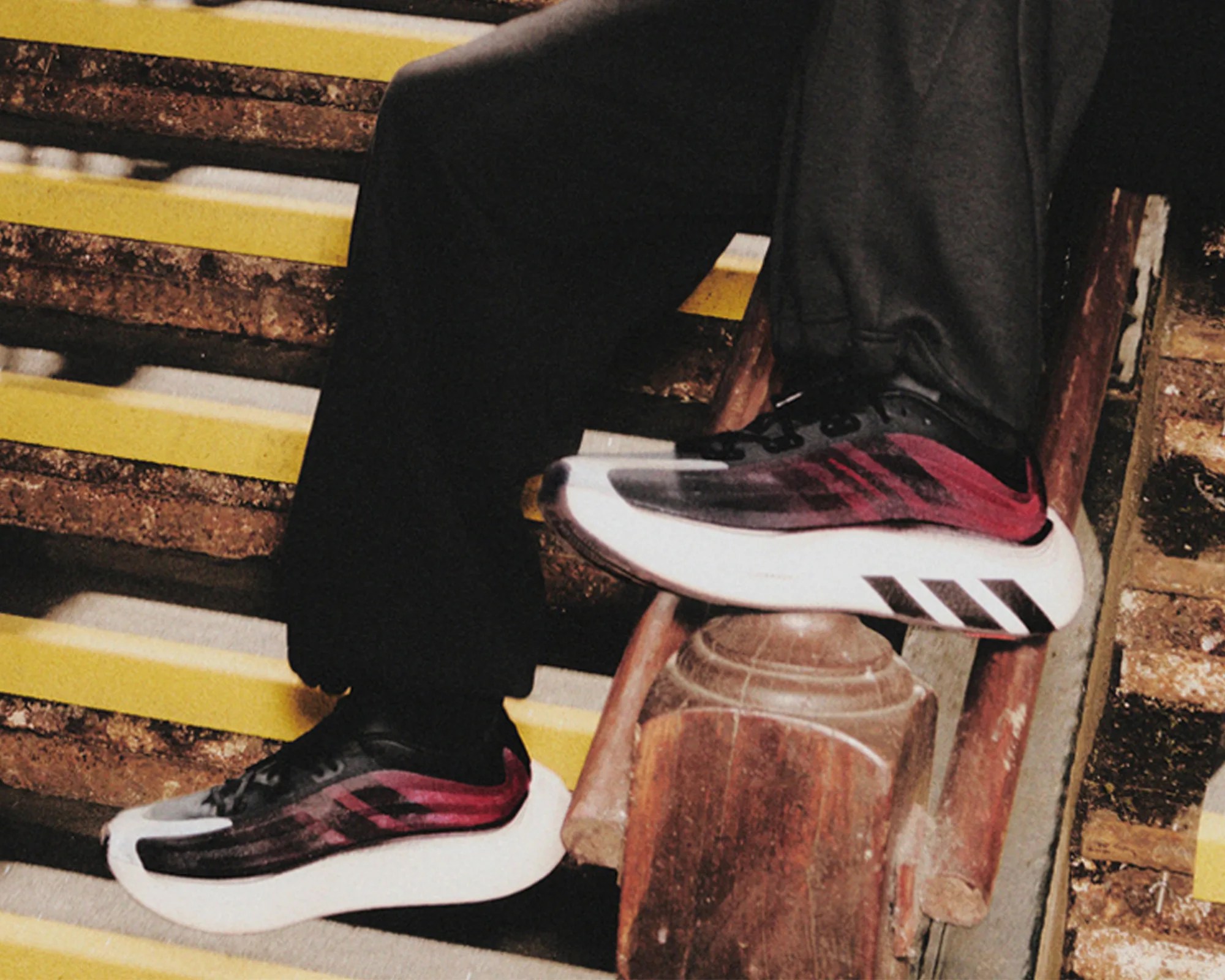 Black and red Adidas running shoes worn with black pants on yellow-striped concrete stairs.