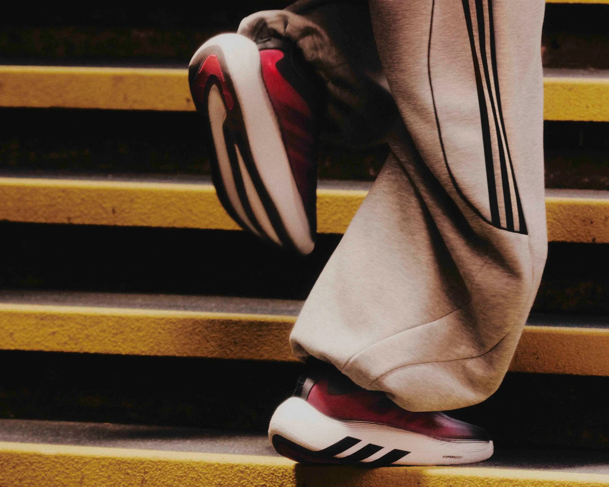 Red and black Adidas sneakers with thick white soles worn with beige pants with black stripes on yellow stairs.