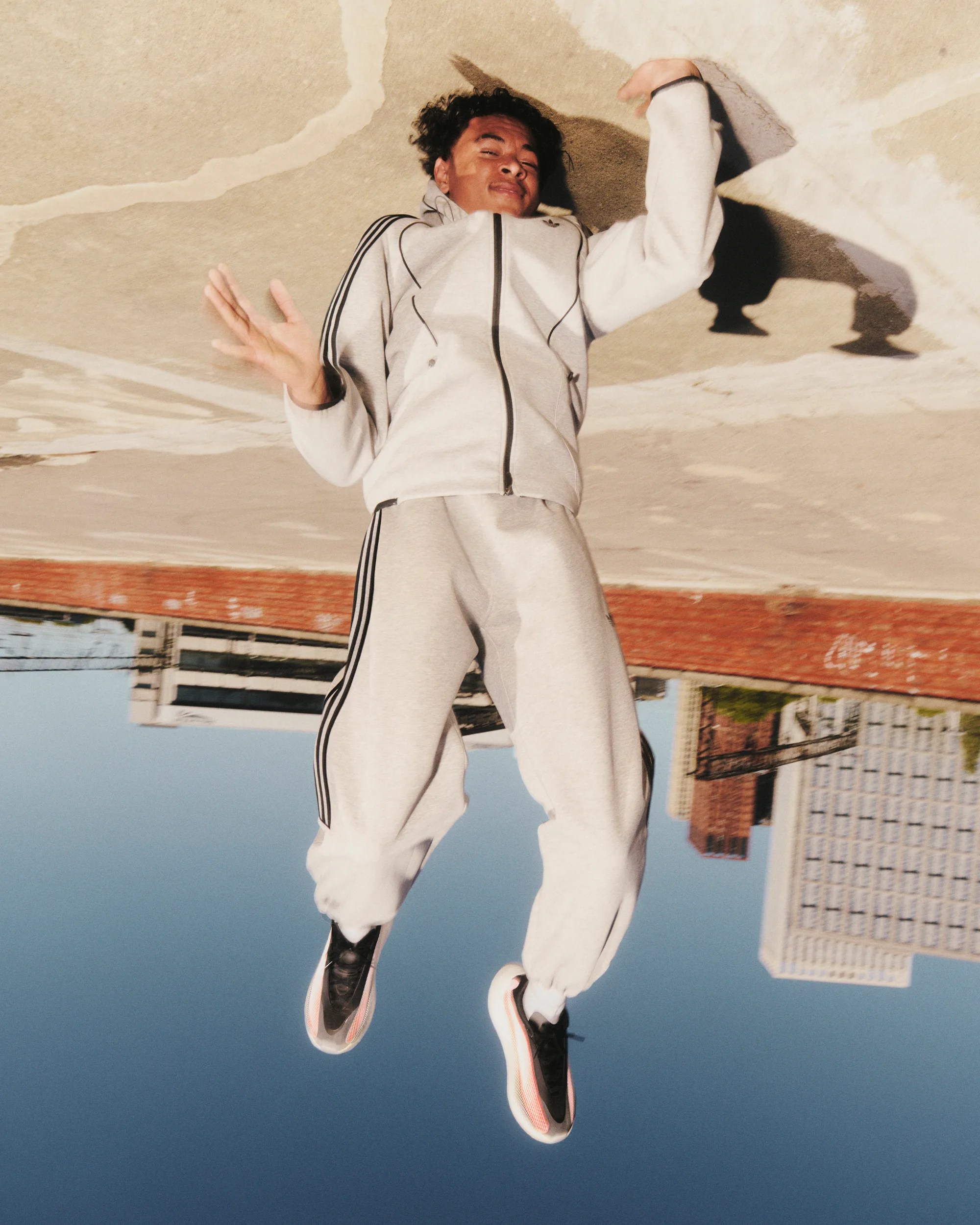 Person in light gray Adidas tracksuit with black stripes jumping outdoors on a sunny day.