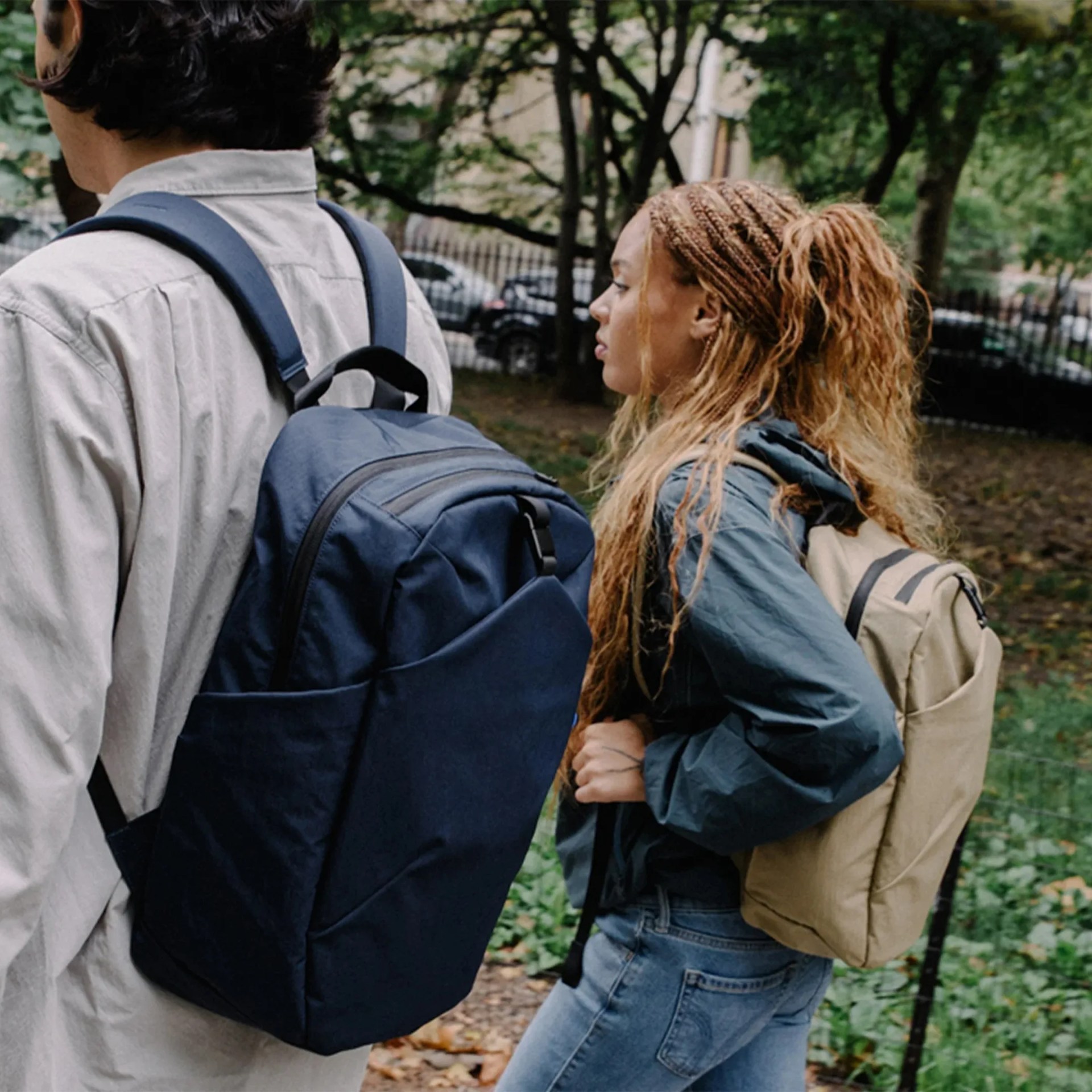 Two people walking outdoors wearing backpacks, one with a navy blue backpack and the other with a beige backpack.