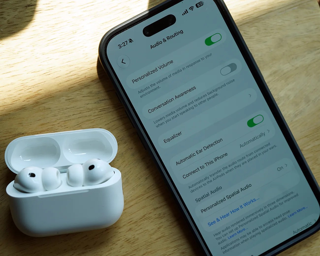 White wireless earbuds in an open charging case next to a smartphone displaying audio and routing settings on a wooden surface.