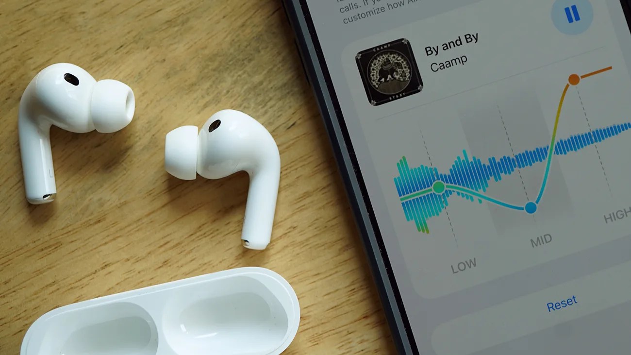 Pair of white wireless earbuds with silicone tips next to an open charging case on a wooden surface, beside a smartphone screen showing an audio equalizer.