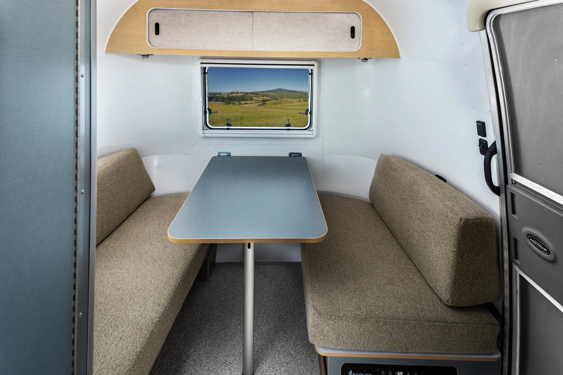 Compact camper dining area with gray table, beige cushioned benches, and a window showing a grassy landscape.