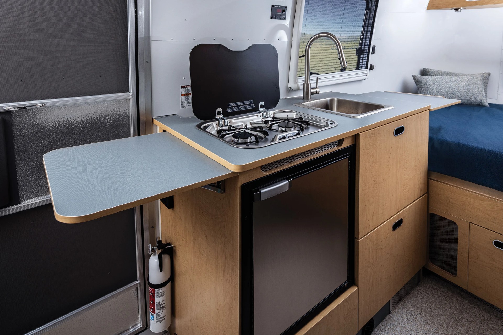 Compact camper kitchen with two-burner stove, stainless steel sink, fold-out countertop extensions, and wooden cabinetry.