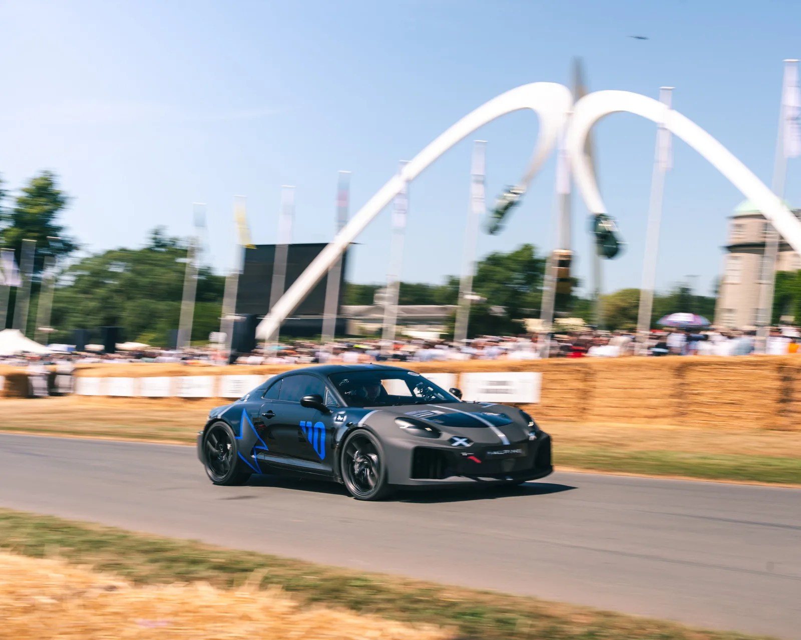Matte black Alpine race car with blue number 70 and star decal driving on a track with hay bales and crowd in background.