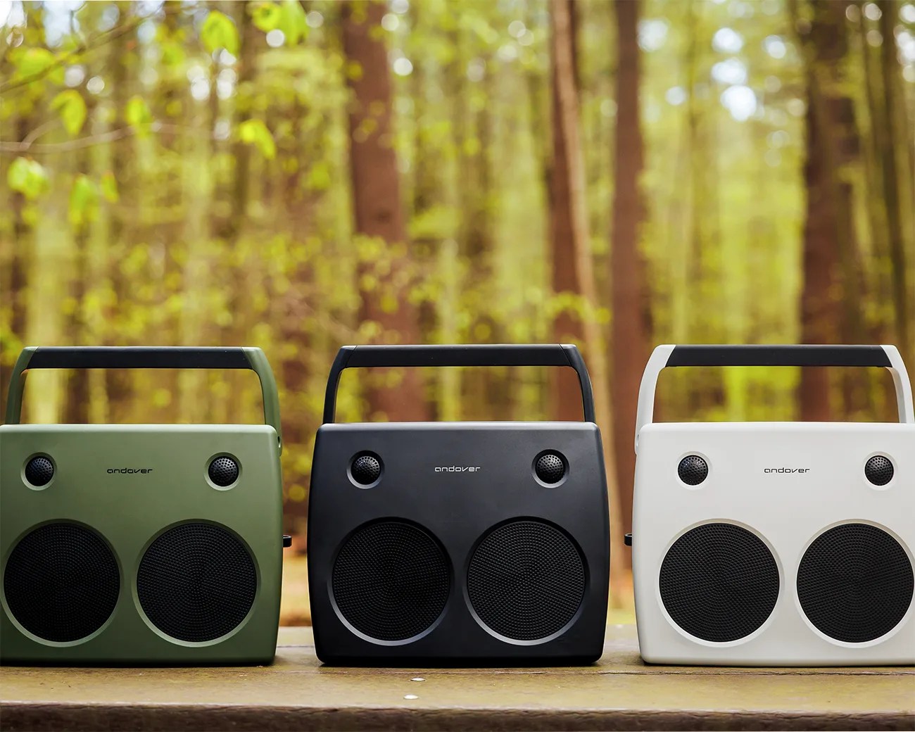 Three portable Andover speakers in green, black, and white with handles, placed on a wooden surface outdoors.