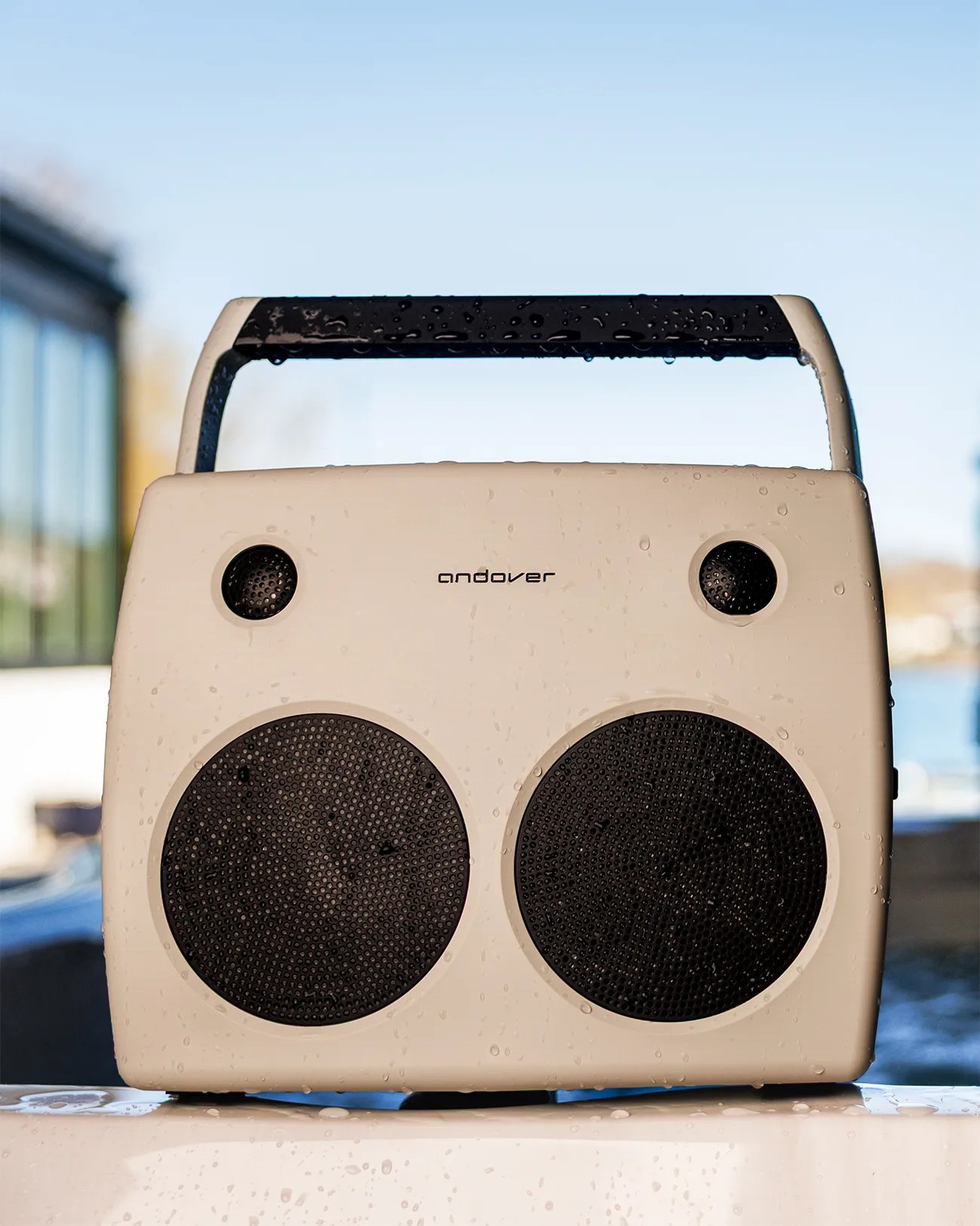 White Andover portable speaker with black handle and four visible speaker grills, covered in water droplets.