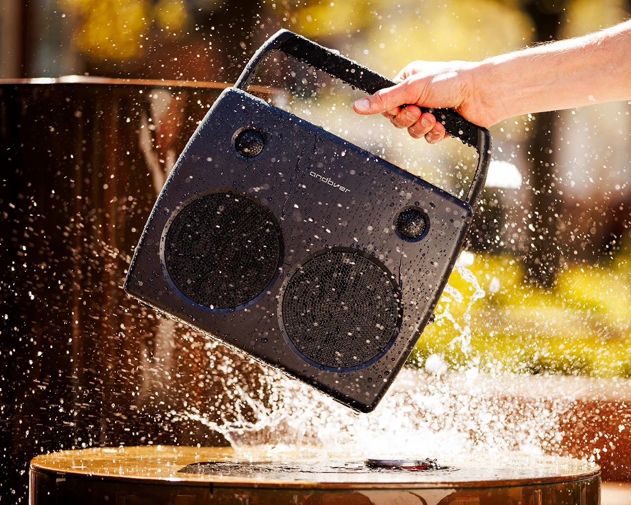 Black portable speaker with handle, wet and splashing water, held in a hand above a wooden surface.