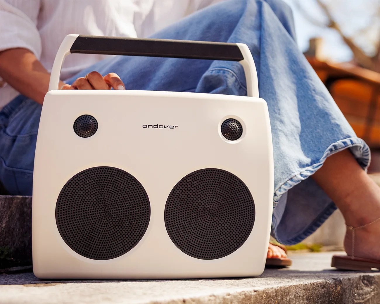 White portable speaker with black mesh grills and a black handle, placed outdoors near a person in blue jeans.