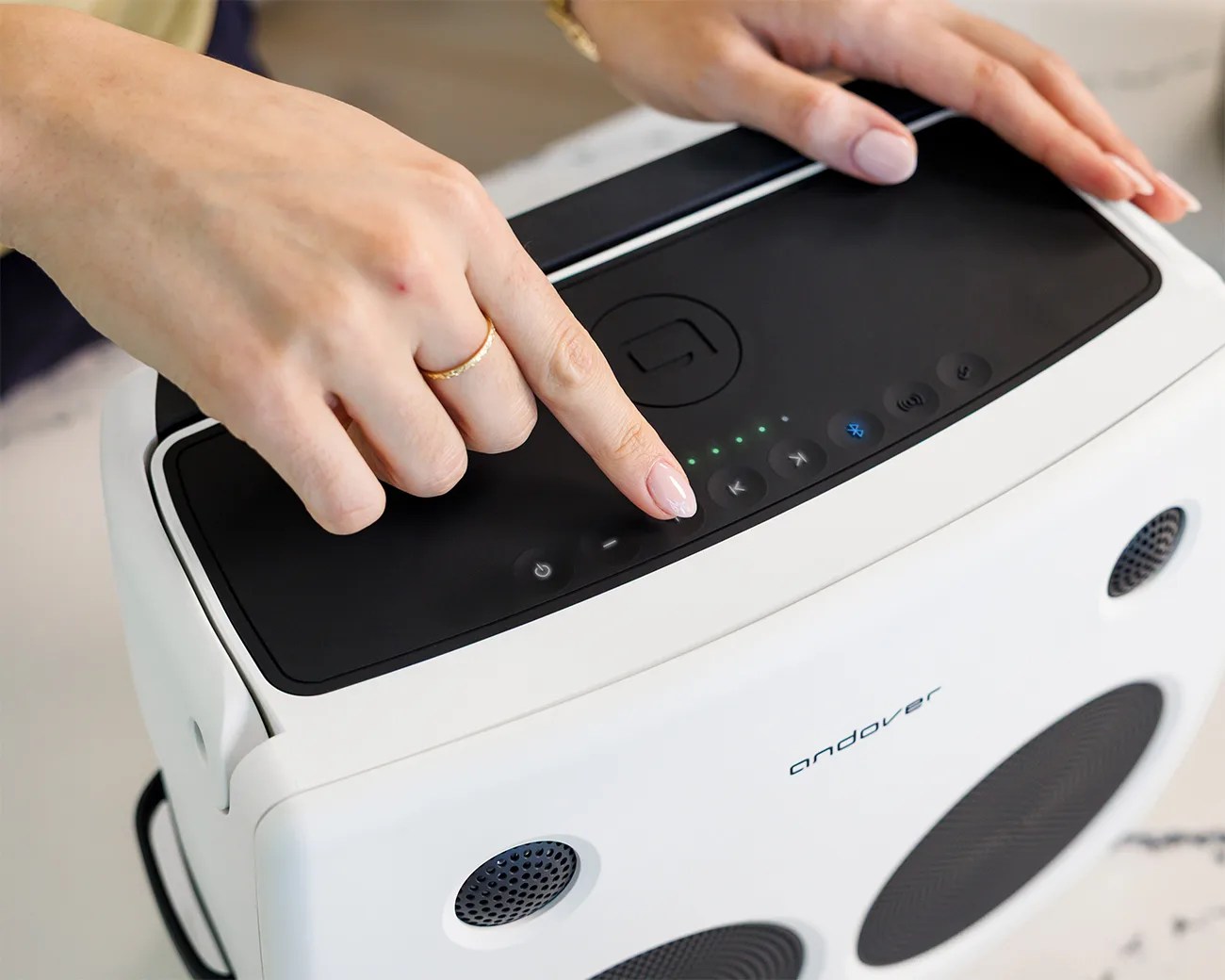 White Andover portable speaker with black control panel and Bluetooth button lit, held by hands with a gold ring.
