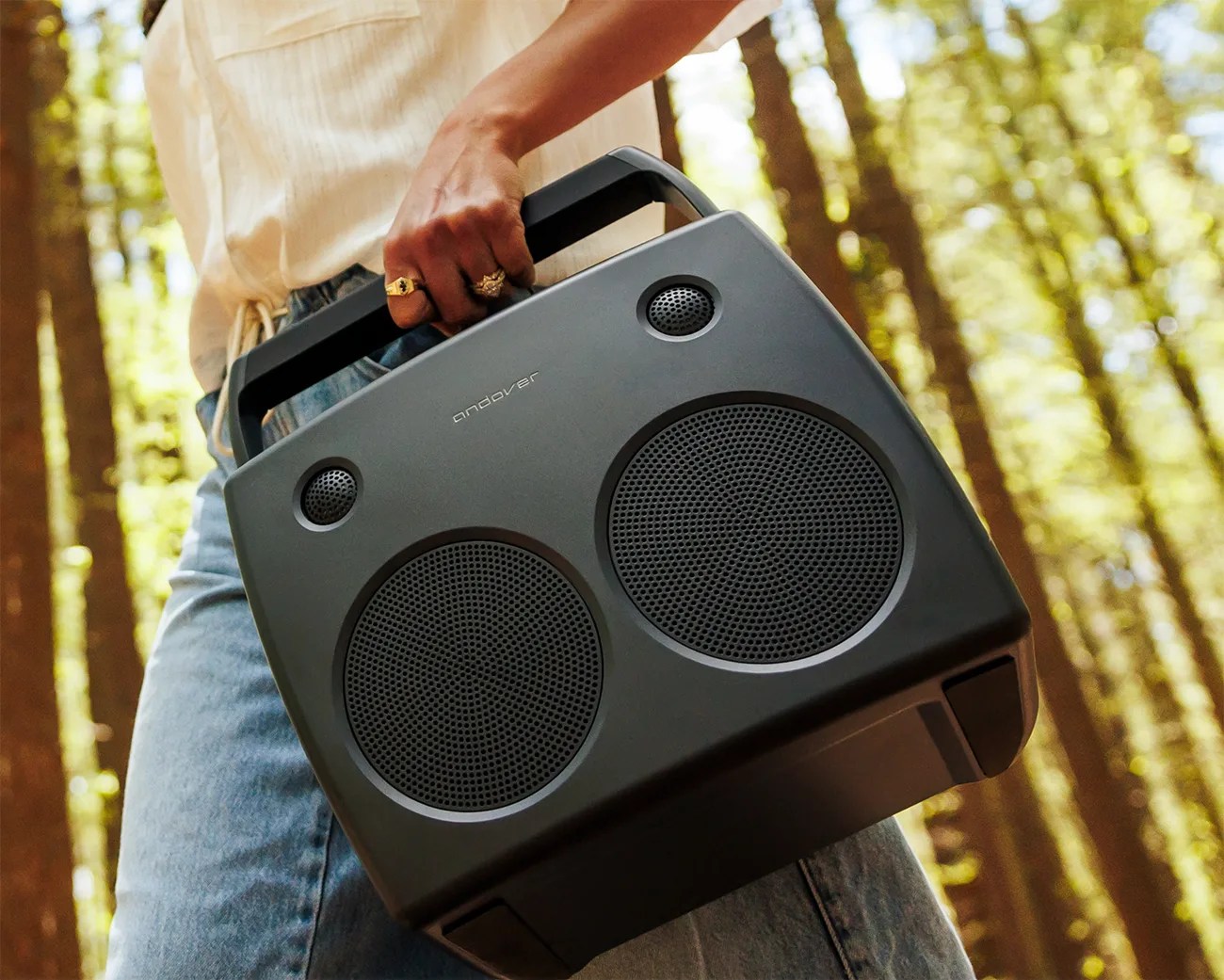 Black portable speaker with two large and two small round speakers held by a person in a forest setting.