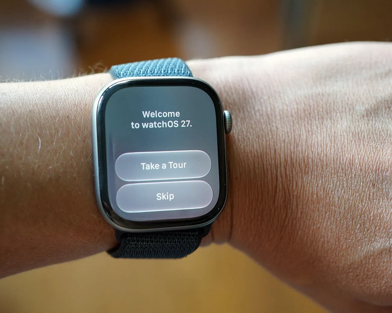 Smartwatch with black case and blue fabric band displaying "Welcome to watchOS 27" screen with "Take a Tour" and "Skip" buttons.