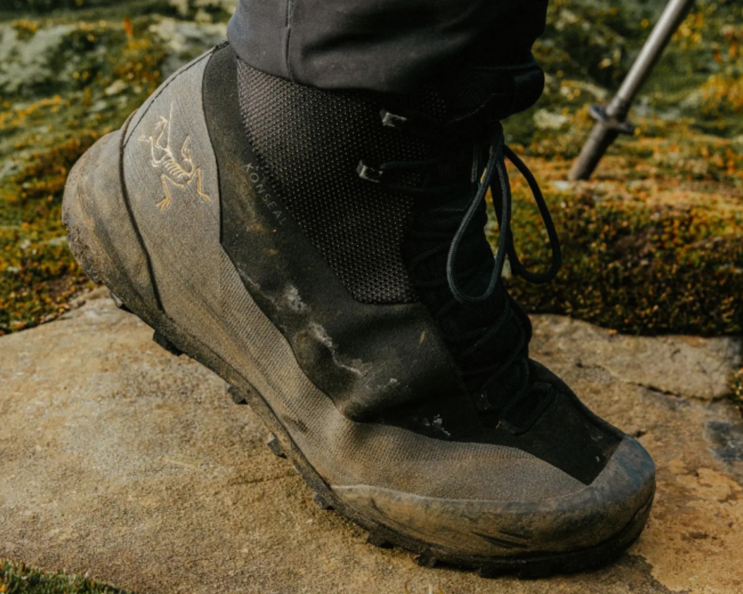 Black and gray hiking boot with laces and a yellow skeletal bird logo on the side, worn on a rocky surface.
