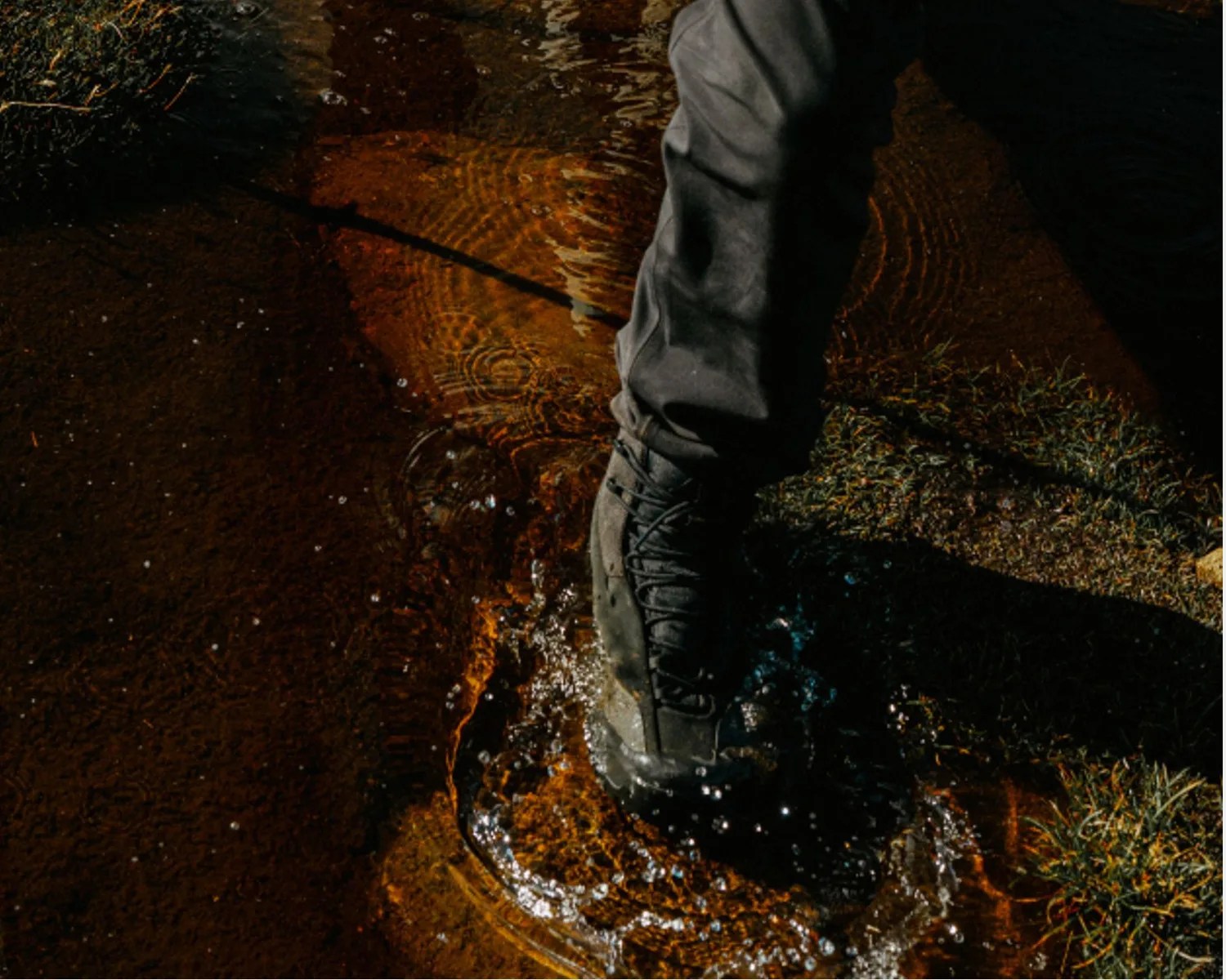 Black hiking boot stepping into shallow water with ripples and splashes.