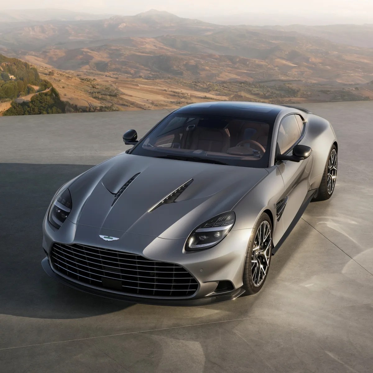 Silver Aston Martin sports car with black roof and large front grille parked on concrete with mountainous landscape in background.