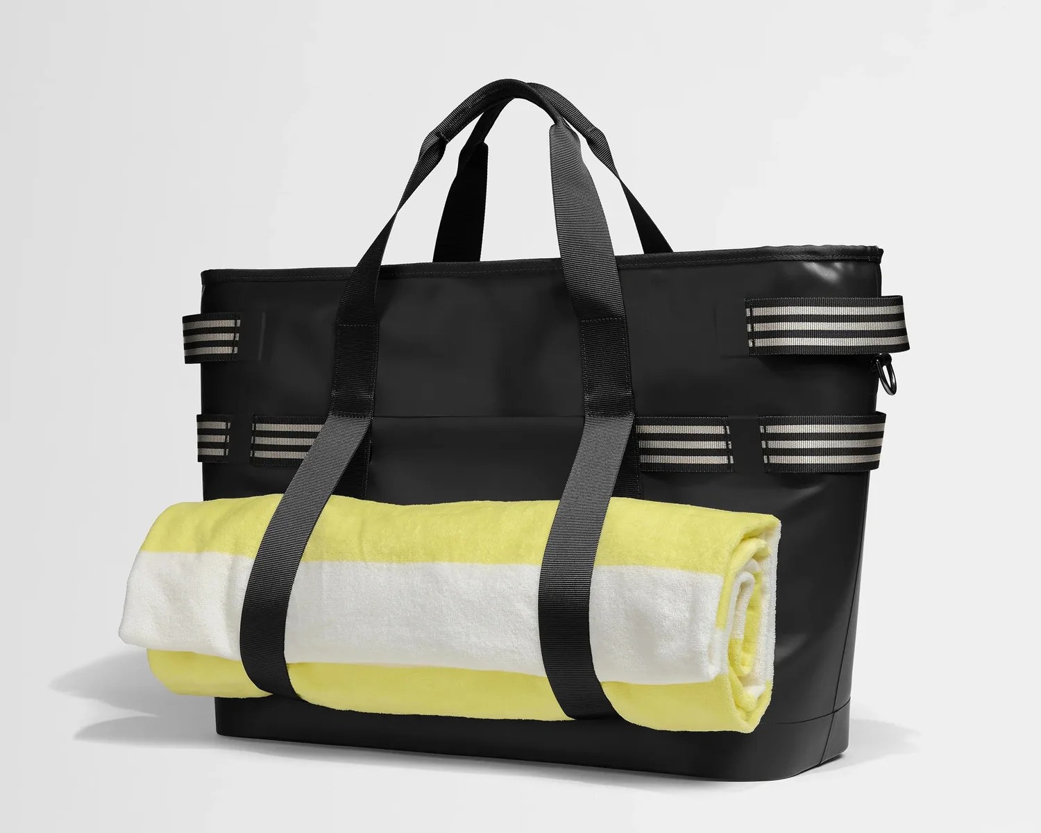 Black tote bag with black and white striped straps holding a rolled yellow and white beach towel.