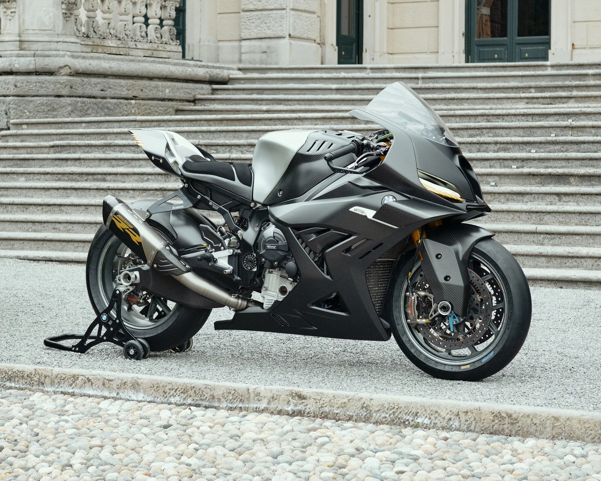Matte black and silver sport motorcycle with carbon fiber body panels parked on gravel in front of stone steps.