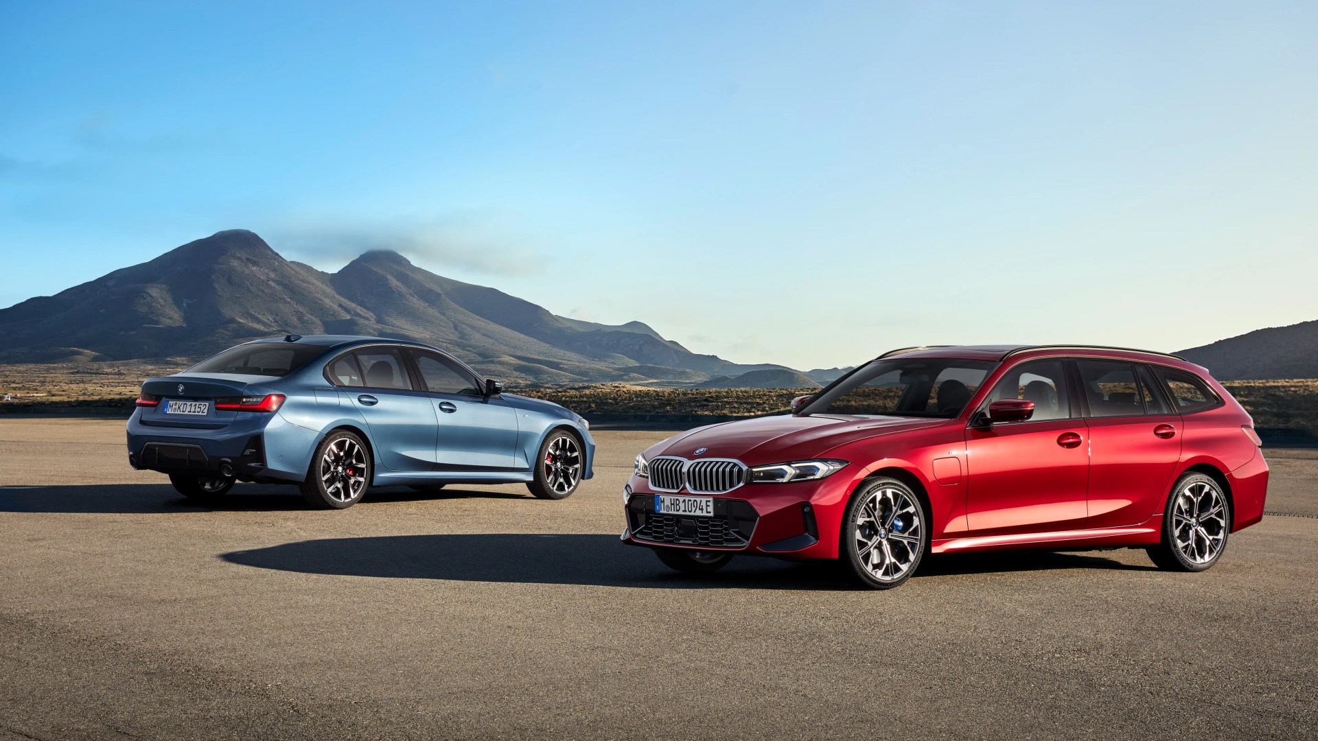 Red BMW station wagon and blue BMW sedan parked on a flat open area with mountains in the background.