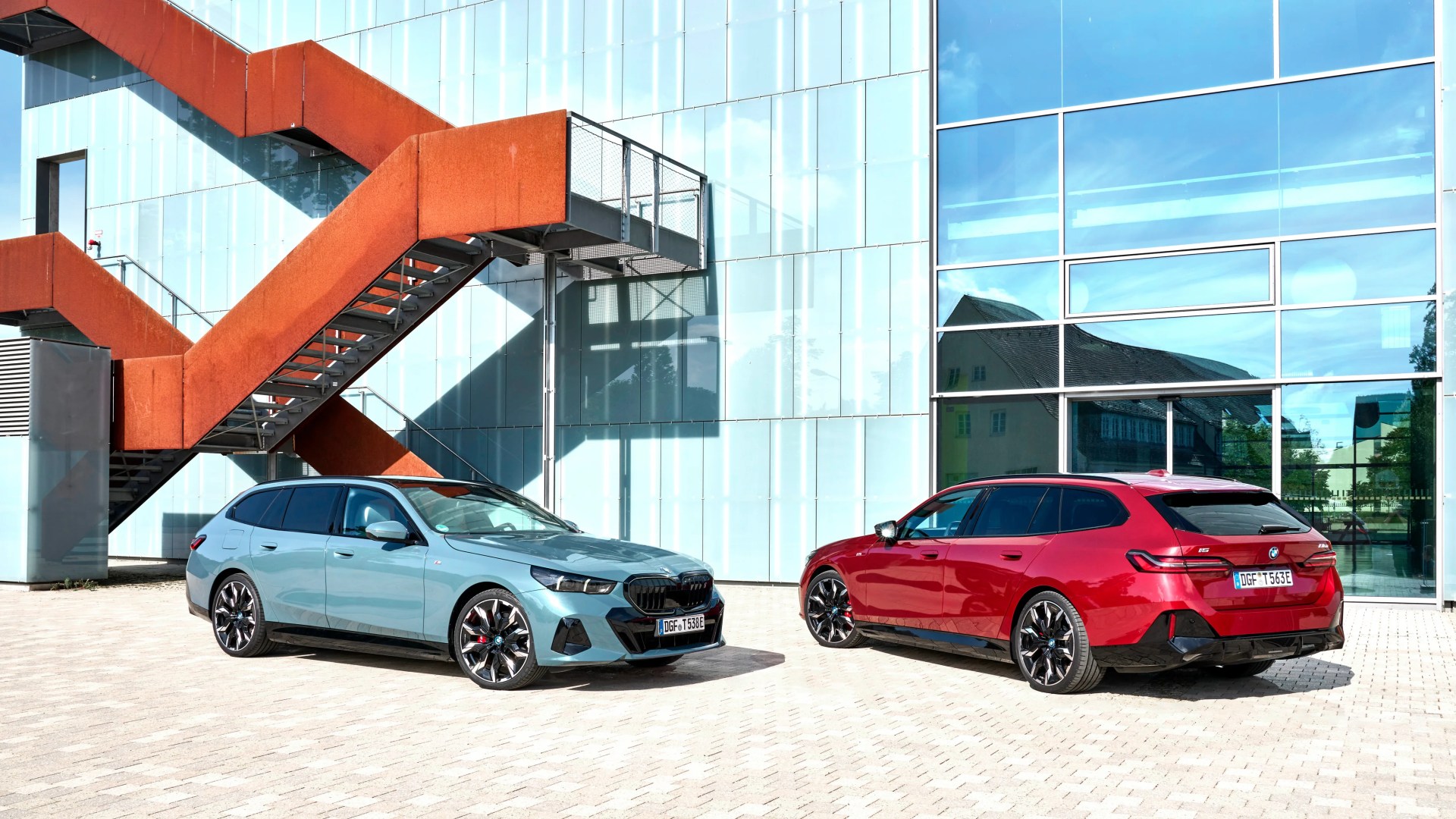 Two BMW station wagons, one light blue and one red, parked in front of a modern glass building with a rust-colored exterior staircase.