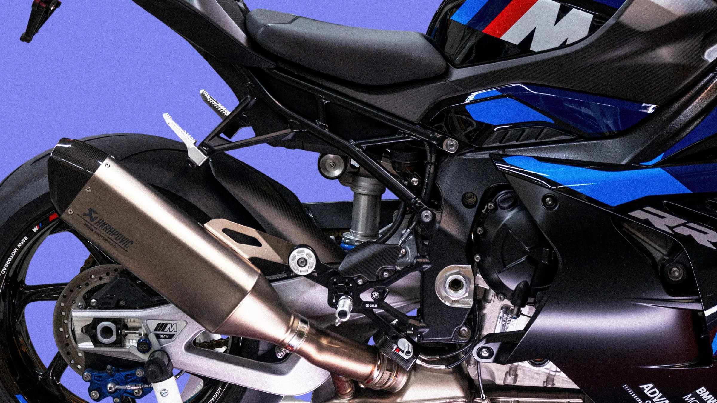 Close-up of a black and blue BMW motorcycle's rear section showing the Akrapovic exhaust, rear tire, and suspension components.